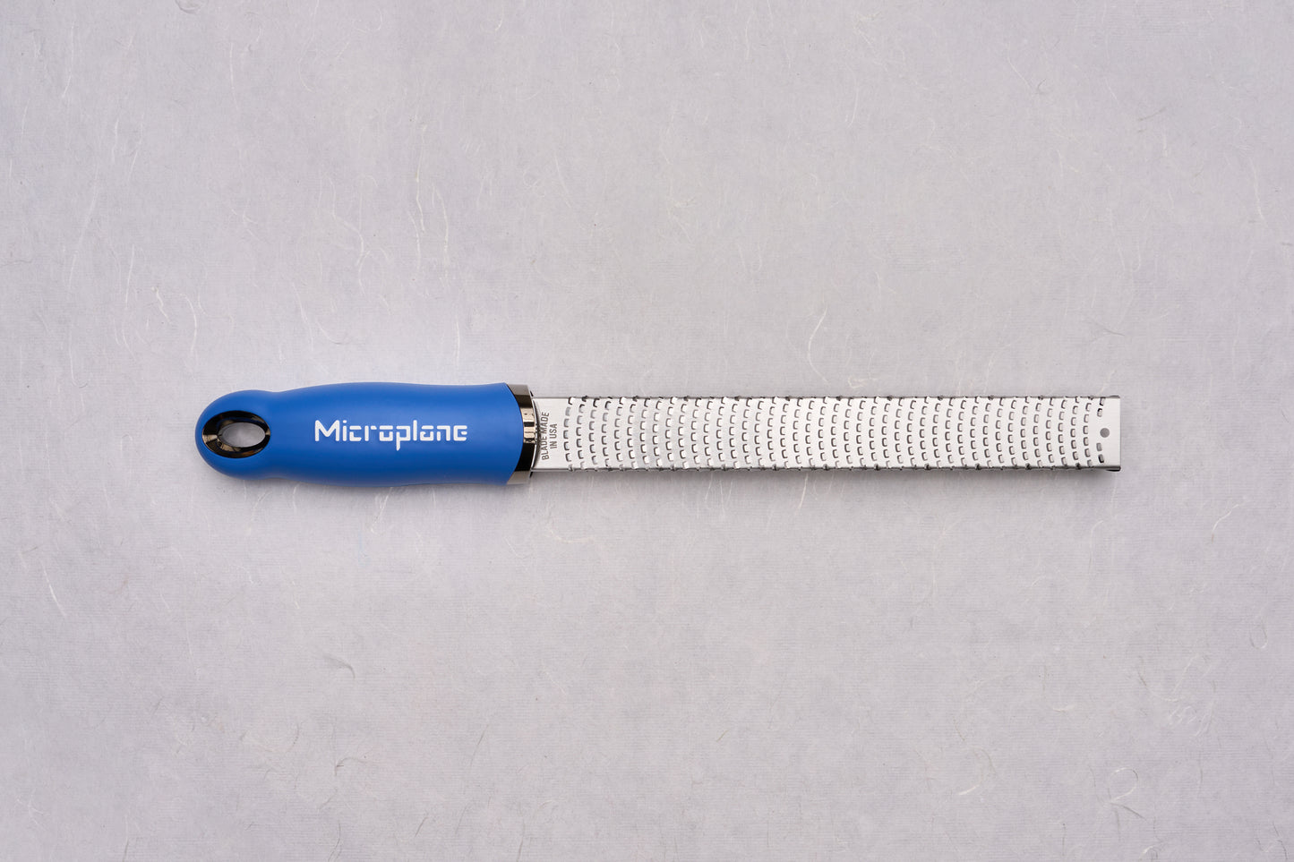 Microplane Premium Classic Zester with blue ergonomic handle and slender stainless-steel grating surface, ideal for zesting citrus and grating hard cheeses, embodies precision craftsmanship cherished by culinary enthusiasts.