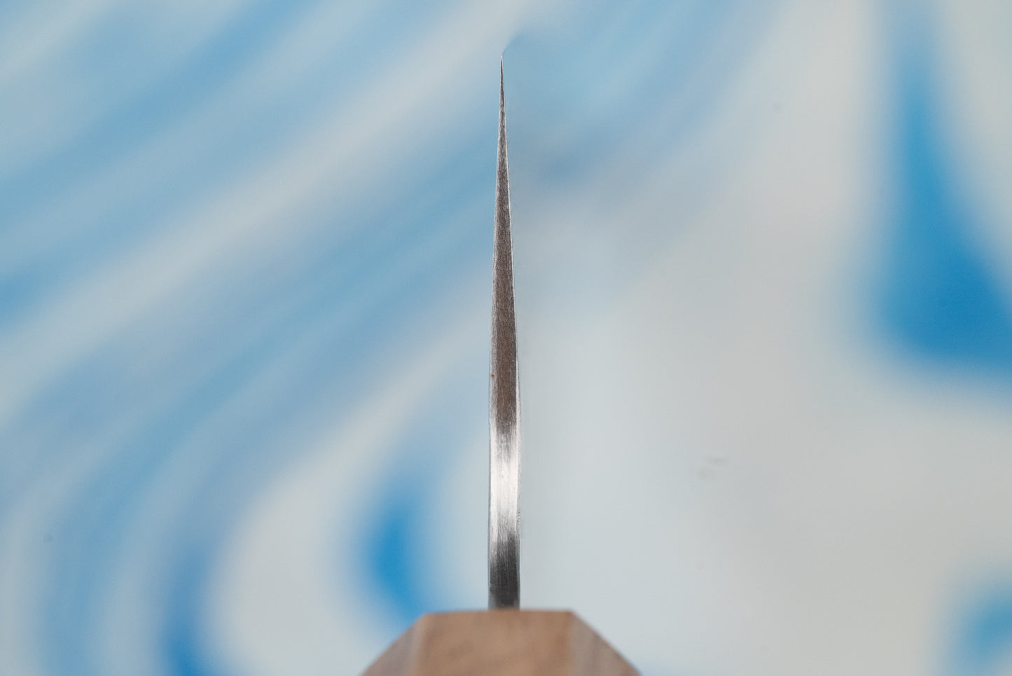 Nigara SPG STRIX Tsuchime Damascus Gyuto 240mm knife blade in profile, showcasing its razor-thin cutting edge and part of the wooden handle against a softly blurred background.