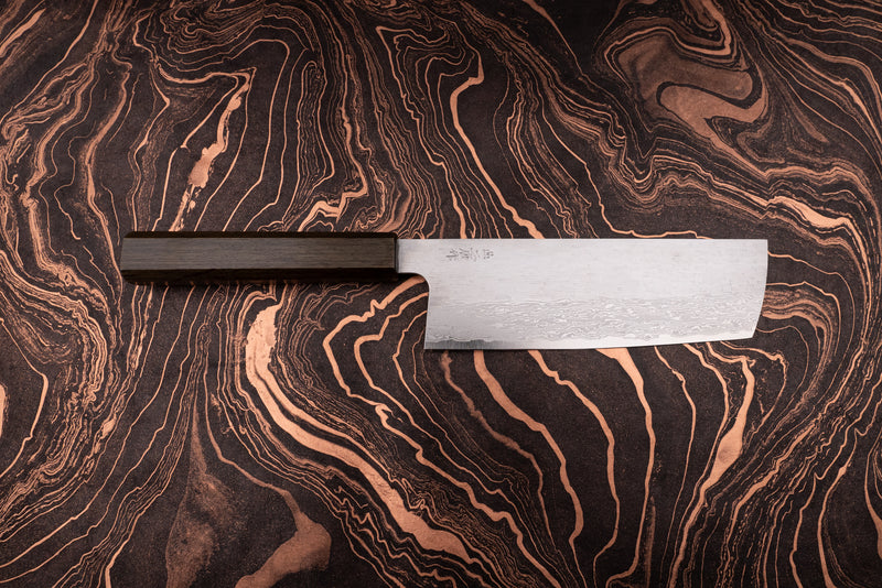 Nigara VG XEOS Tsuchime Damascus Nakiri 180mm, featuring a straight-edged Damascus-pattern blade and octagonal dark wooden handle, displayed on a marbled coppery surface. Ideal for efficient vegetable chopping.