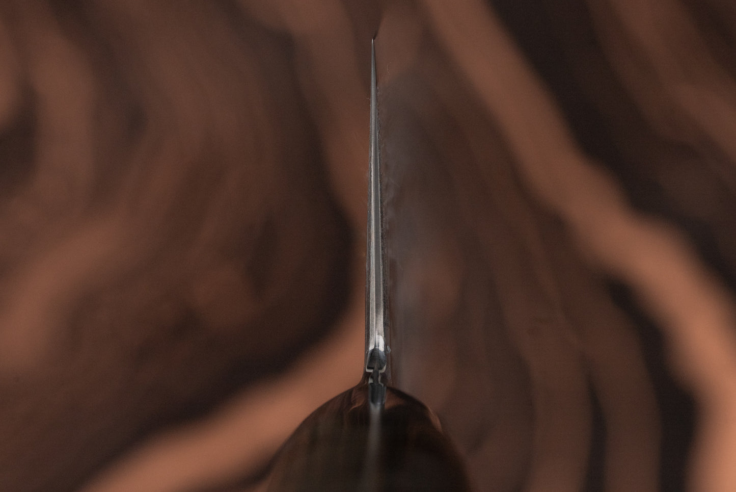 Close-up of the spine of a Ryusen Oukokuryu Gyuto 210mm blade, featuring a glimpse of the wooden handle against a blurred wood-grain background, highlighting its precision craftsmanship and sleek design.