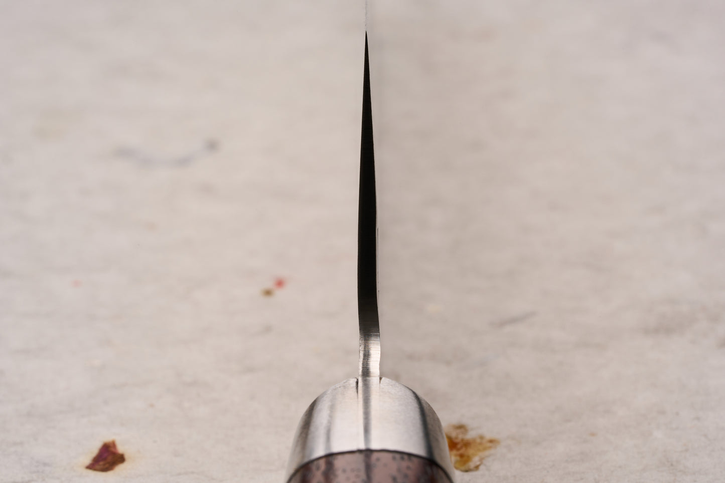 Sakai Takayuki SG2 Tsuchime Damascus Western Kengata Santoku 160mm knife stands upright, showcasing its narrow steel spine and metal bolster, with a glimpse of the wooden handle, on a cloth-dotted surface.
