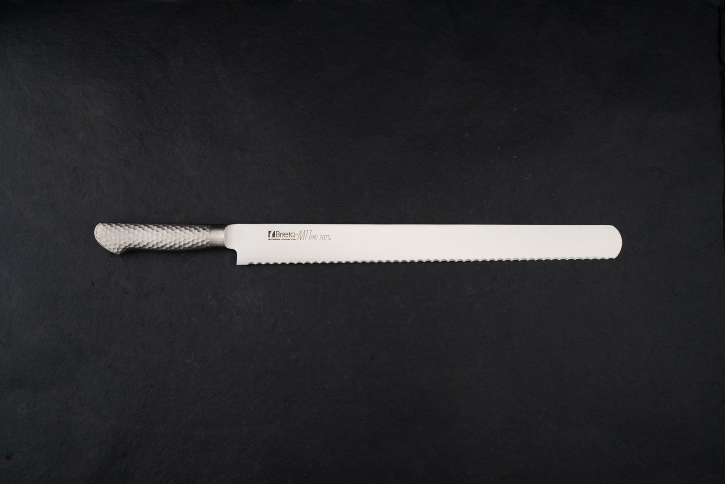 Brieto Pro Bread Knife 390mm M1148 with a long, thin serrated blade and hammered-metal handle, expertly designed for slicing bread without damaging its texture, displayed on a dark background.