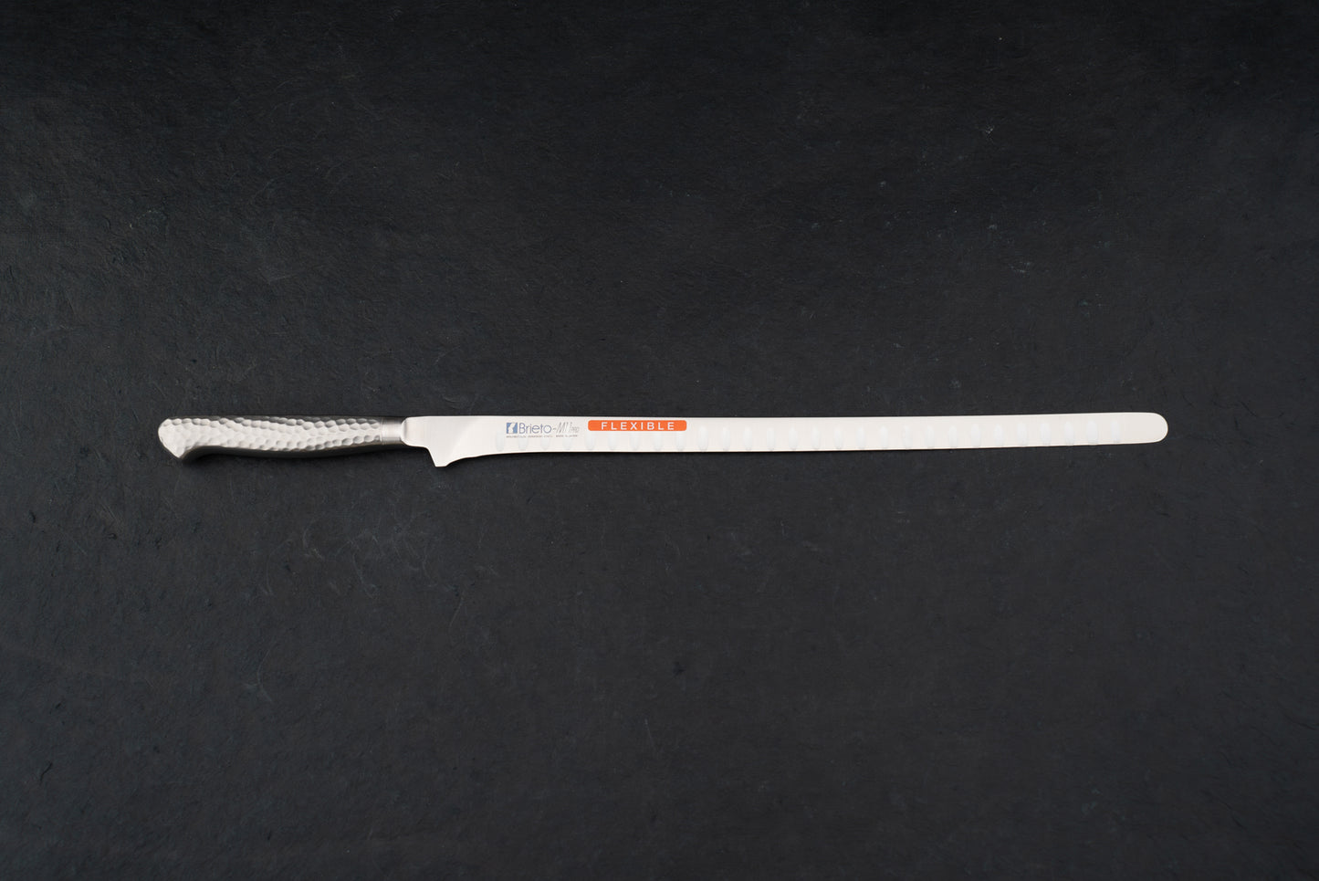 Brieto Pro Flexible Salmon Slicer with Dimples 320mm M116, featuring a long, narrow, flexible blade and hammered-finish metal handle, ideal for precise slicing of meats and fish, displayed on a dark surface.
