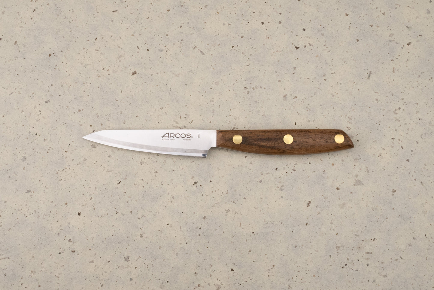 Arcos Nordika 100mm Petty Knife with a straight stainless-steel blade and dark wood handle, lying on a countertop. The blade features the Arcos logo and Made in Spain, Albacete engraving.