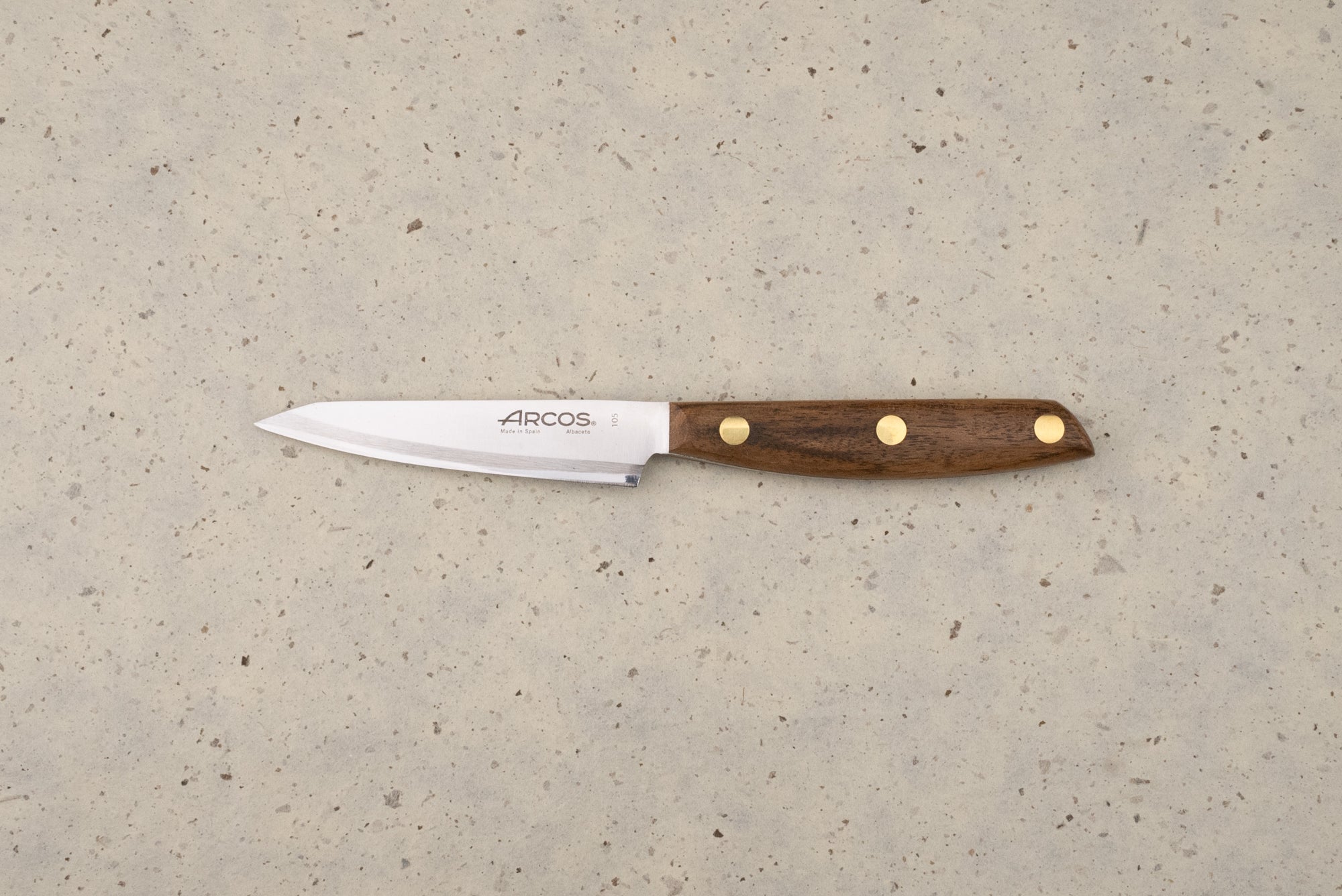 Arcos Nordika 100mm Petty Knife with a straight stainless-steel blade and dark wood handle, lying on a countertop. The blade features the Arcos logo and Made in Spain, Albacete engraving.