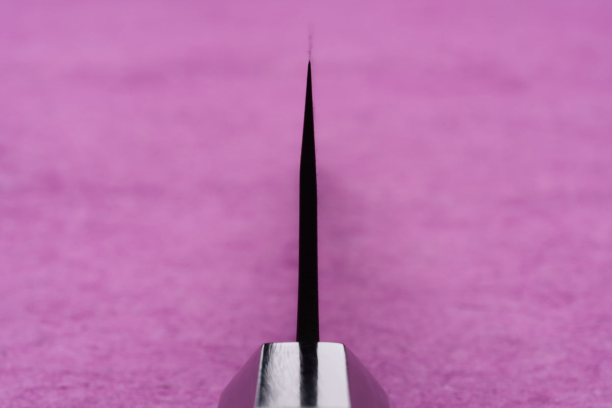 Fujimoto AS Sazanami Gyuto 210mm knife's thin metal spine rests on a pink surface, showcasing its precision-crafted, sharp design.