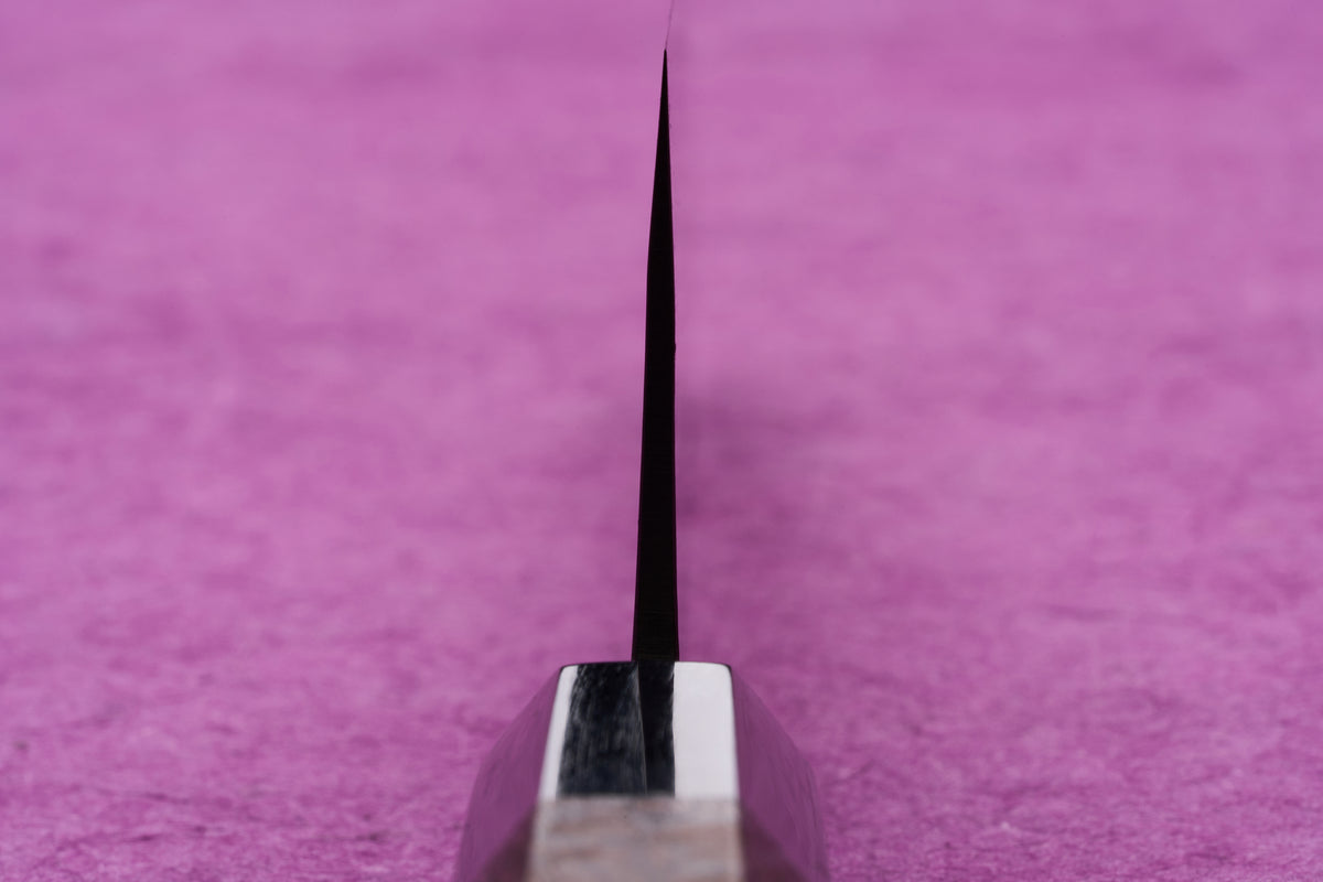 Fujimoto AS Sazanami Santoku 165mm knife's sharp edge and bolster, showcasing its precision craftsmanship and premium materials against a pink background.