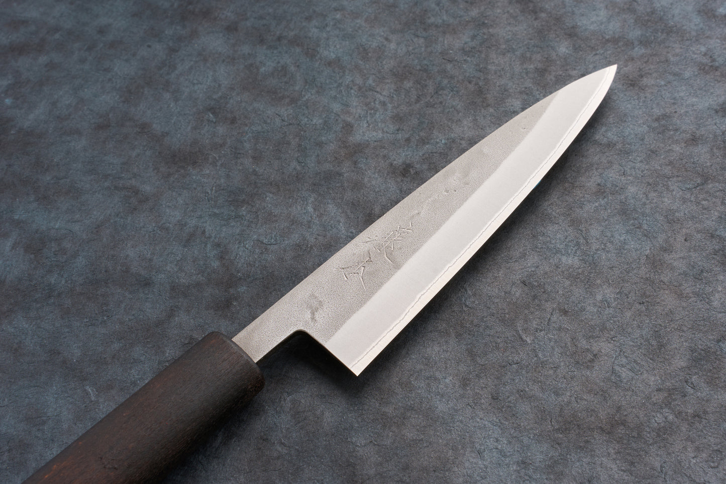 Fujimoto Nashiji Petty 135mm knife, featuring a single-bevel, grain-textured steel blade with stamped Japanese characters, and a dark wa-style wooden handle, ideal for precise cutting tasks on a board.