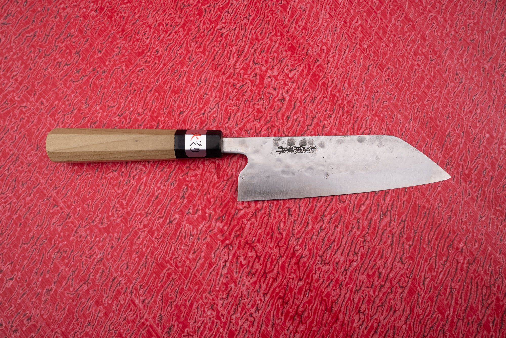 Fujiwara Maboroshi Wa Santoku 165mm knife features a hammered-finish straight-edged carbon steel blade with etched Japanese characters and an octagonal light wood handle, lying on a textured red surface.