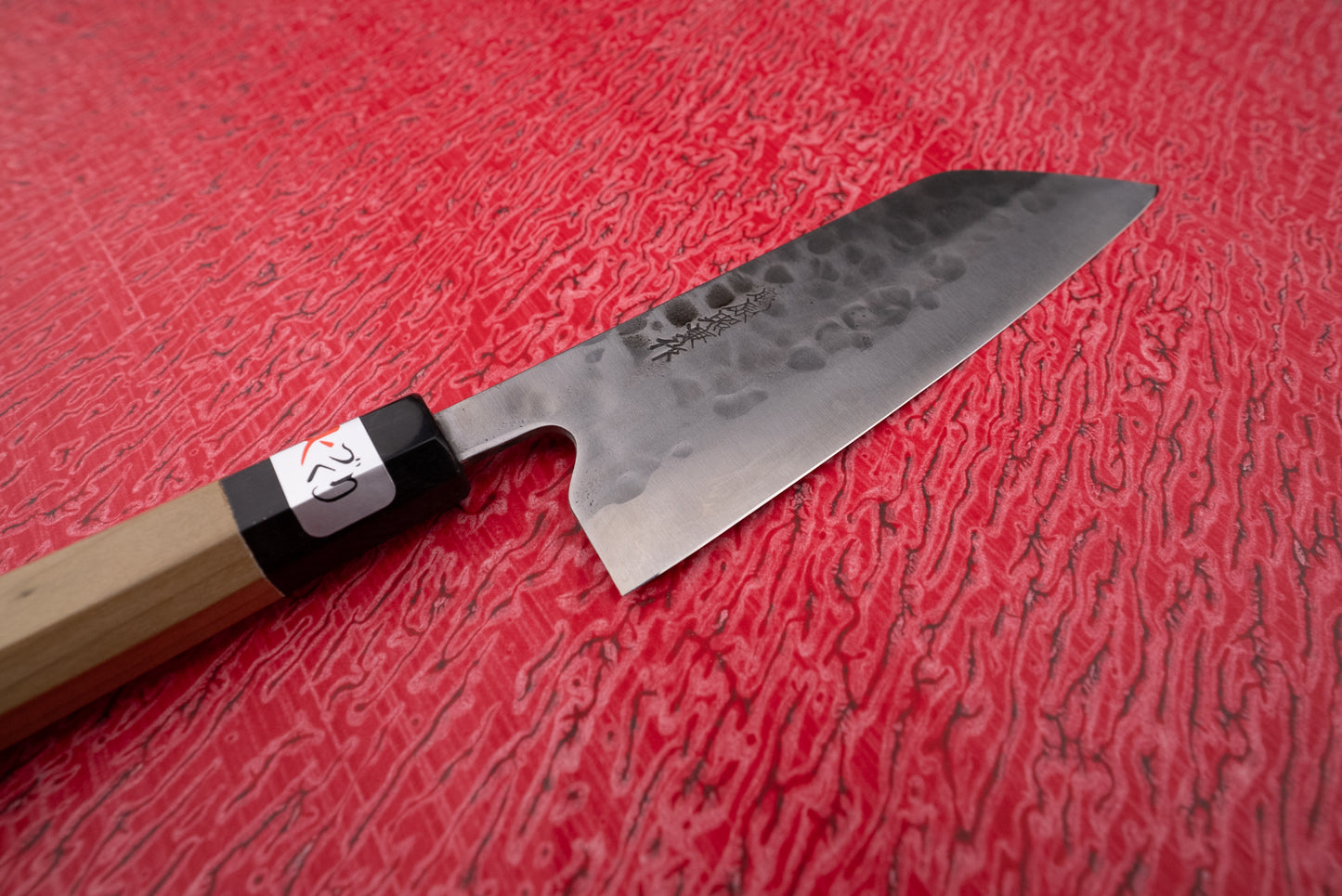 Fujiwara Maboroshi Wa Santoku 165mm, a Japanese-style kitchen knife with a hammered blade and octagonal wooden handle, rests on a red textured cutting board, embodying precision craftsmanship.