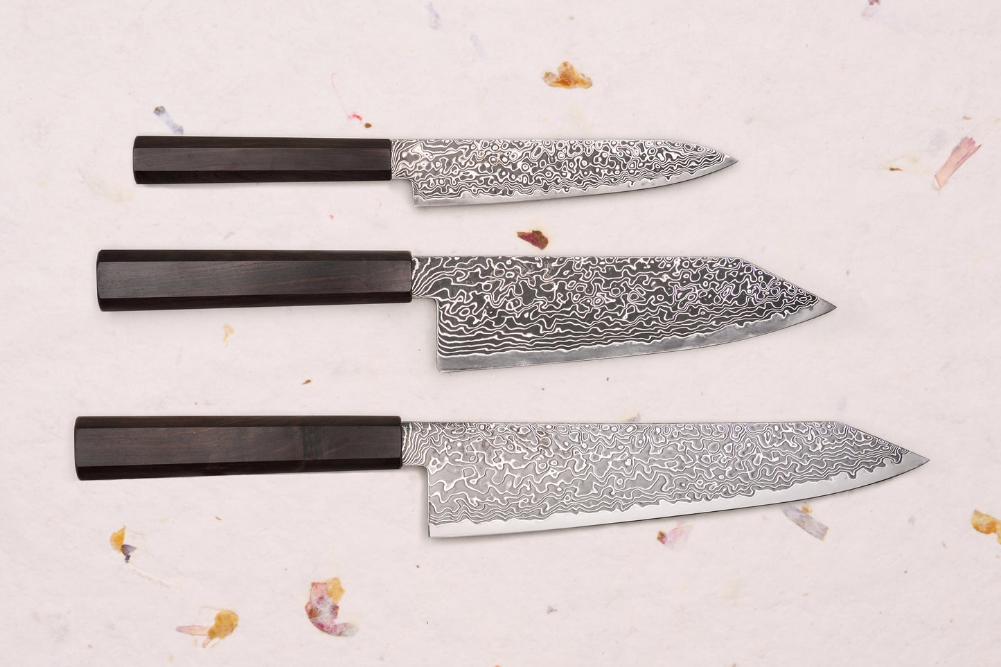 Hatsukokoro Ryuhyo SG2 Kurozome 3 Piece Knife Set with Damascus blades and octagonal wooden handles, displayed on a textured surface with dried floral accents.