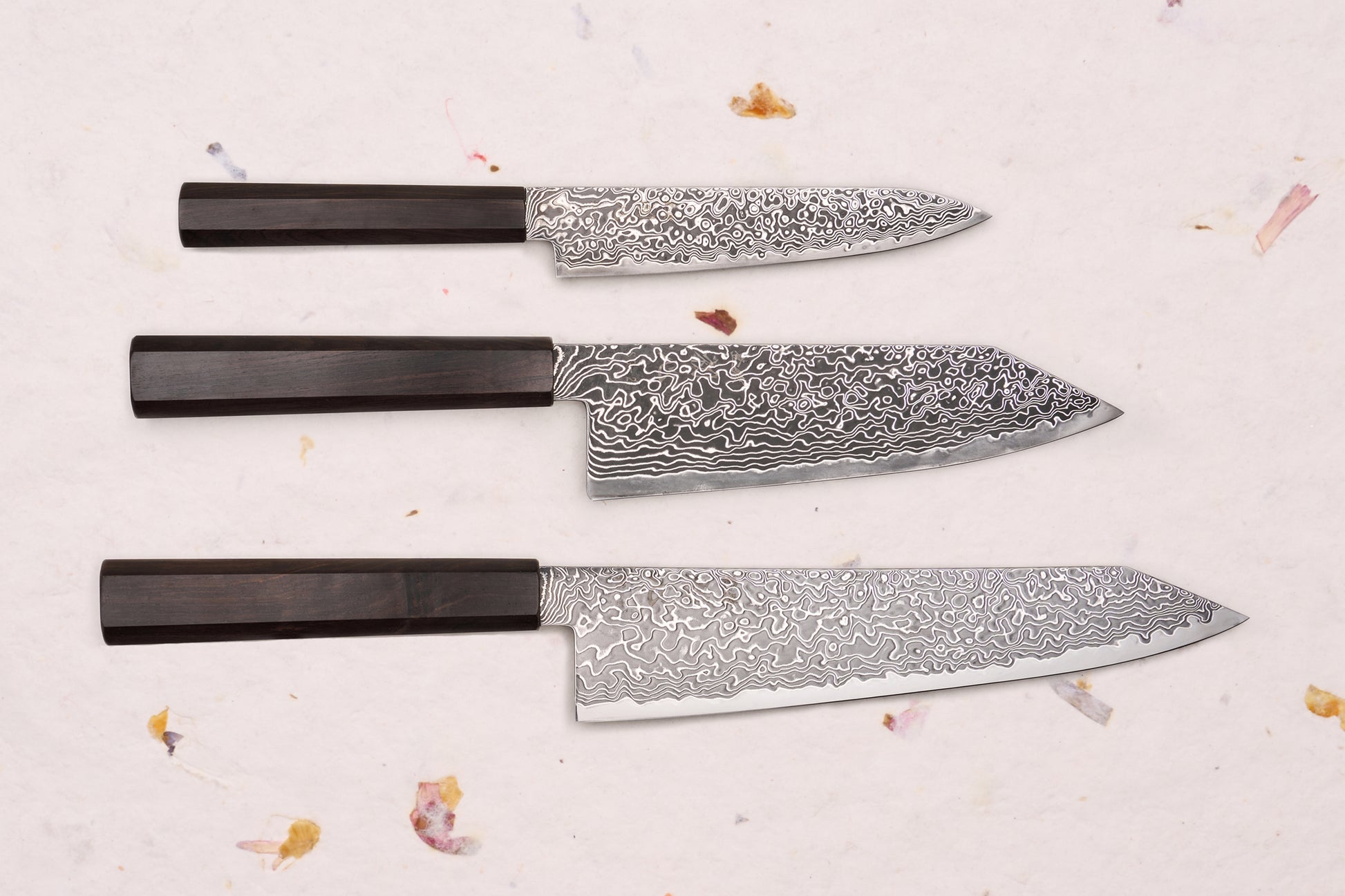 Hatsukokoro Ryuhyo SG2 Kurozome 3 Piece Knife Set with Damascus blades and octagonal wooden handles, displayed on a textured surface with dried floral accents.