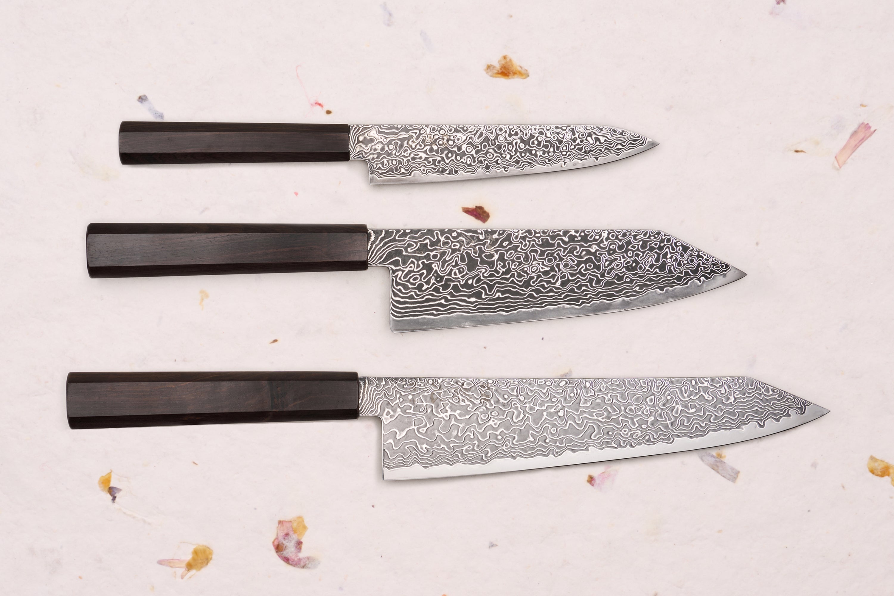 Hatsukokoro Ryuhyo SG2 Kurozome 3 Piece Knife Set with Damascus blades and octagonal wooden handles, displayed on a textured surface with dried floral accents.