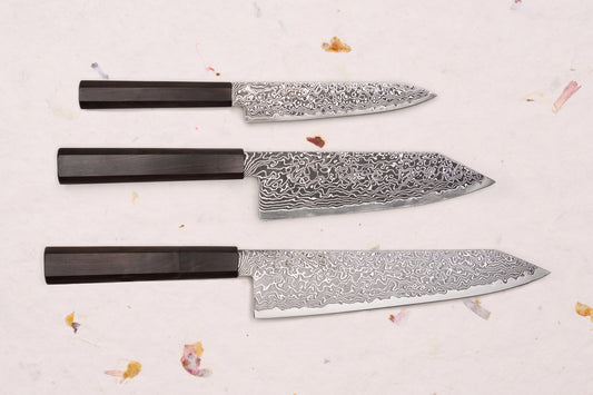 Hatsukokoro Ryuhyo SG2 Kurozome 3 Piece Knife Set with Damascus blades and octagonal wooden handles, displayed on a textured surface with dried floral accents.