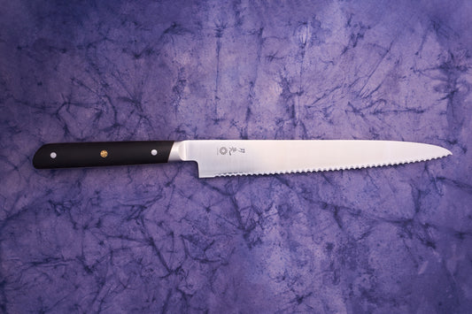 Hazaki Classic Black Bread Knife 240mm features a polished scalloped steel blade with a black handle and brass emblem, showcasing Japanese craftsmanship on a purple marbled fabric backdrop.