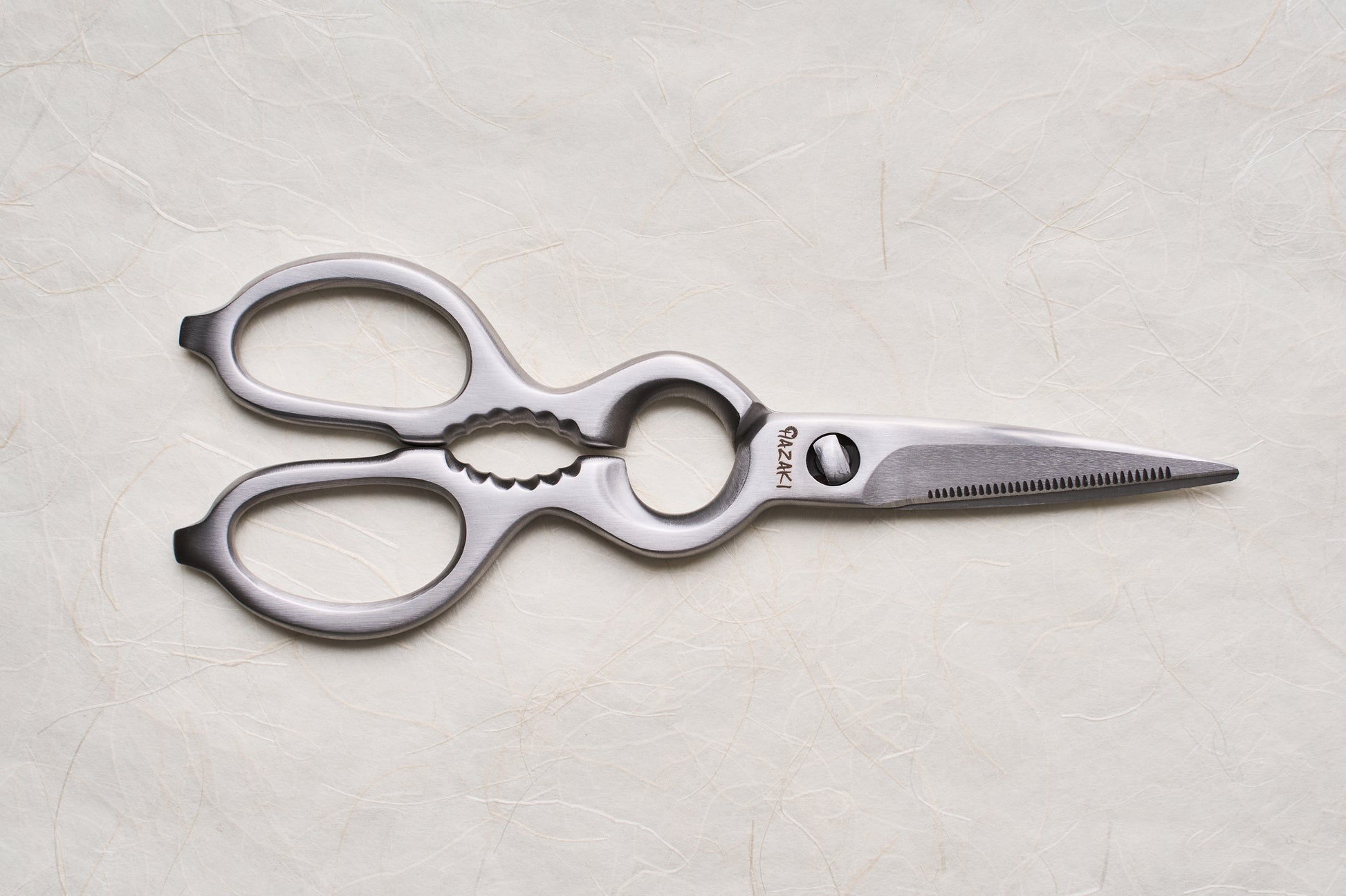 Hazaki Stainless Kitchen Shears with ergonomic handles and a serrated blade, featuring a bottle opener and nutcracker, displayed on a textured surface. Ideal for versatile kitchen tasks and easy maintenance.