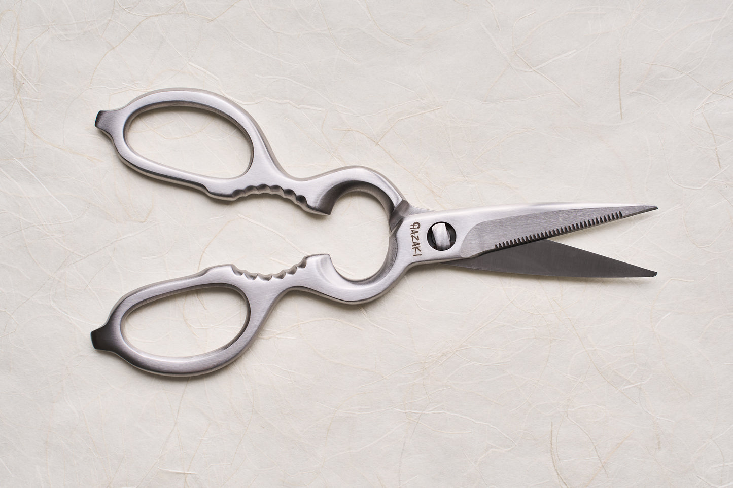 Hazaki Stainless Kitchen Shears with serrated blades and oversized metal handles, featuring a nutcracker and bottle opener, lie slightly open, showcasing their ergonomic and fully separable design for easy cleaning and sharpening.