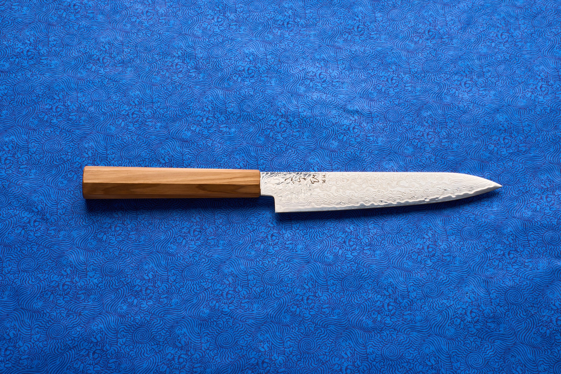 ICHIGO Haku Petty 150mm knife with a Damascus-pattern blade and a straight wooden handle, displayed on a blue patterned surface, ideal for precise cutting tasks like slicing shallots and herbs.