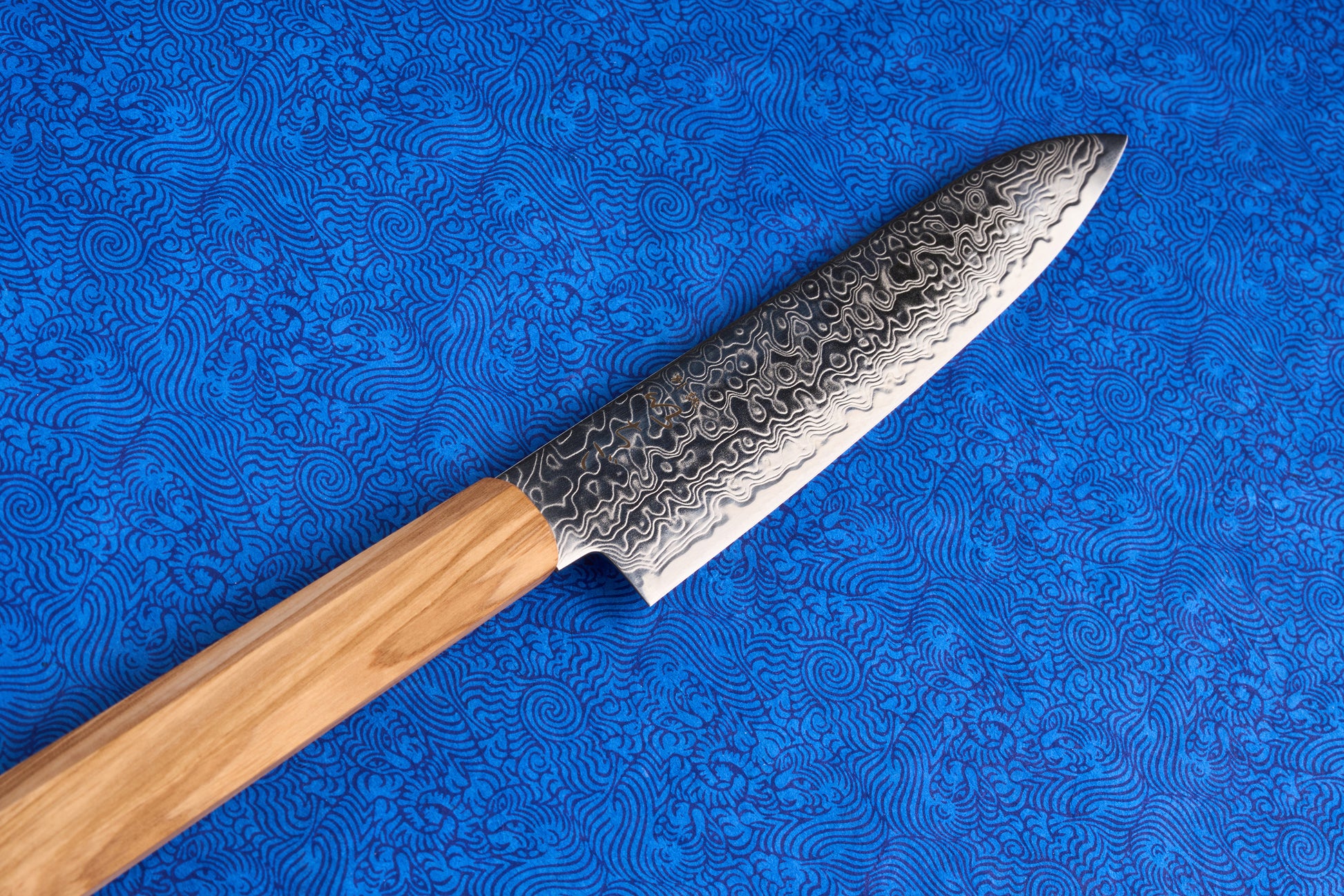 ICHIGO Haku Petty 150mm knife with a Damascus-steel blade, showcasing its wavy pattern, rests diagonally on a blue cloth with abstract floral motifs, emphasizing its elegance and precision for smaller kitchen tasks.