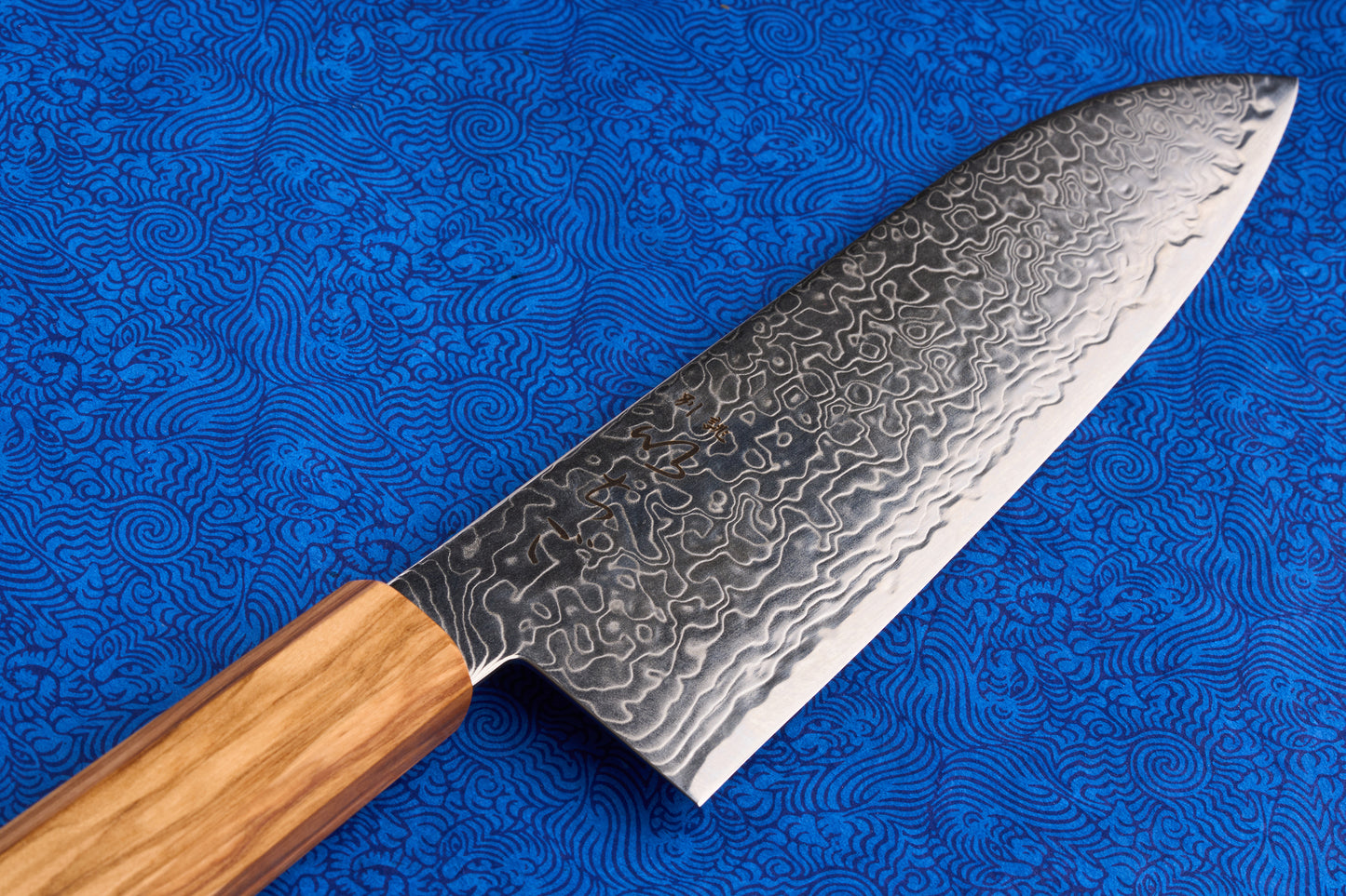 ICHIGO Haku Santoku 180mm features a patterned Damascus steel blade and a light-wood handle, perfect for multi-purpose kitchen tasks, set against a blue swirled fabric background.