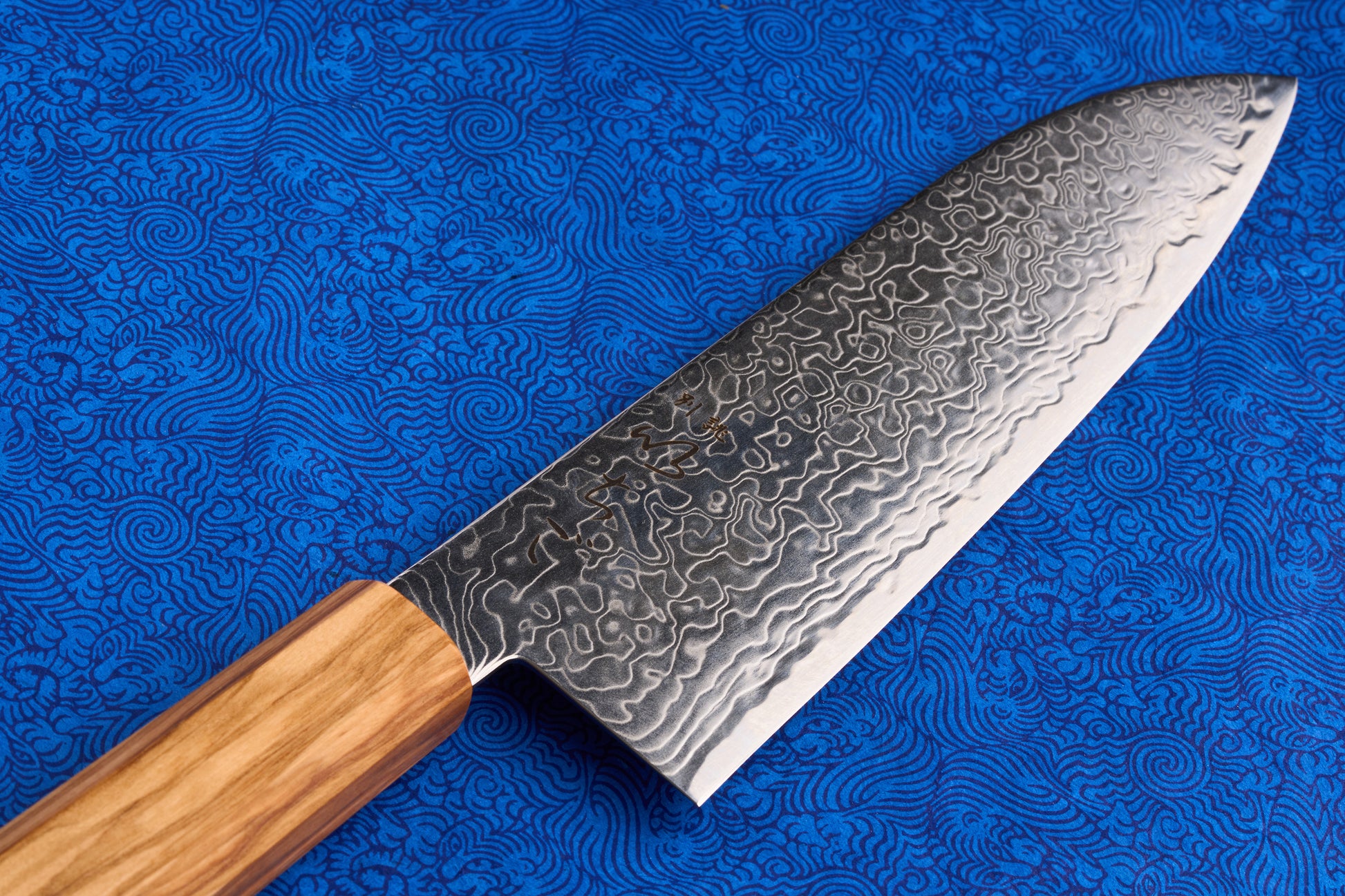 ICHIGO Haku Santoku 180mm features a patterned Damascus steel blade and a light-wood handle, perfect for multi-purpose kitchen tasks, set against a blue swirled fabric background.