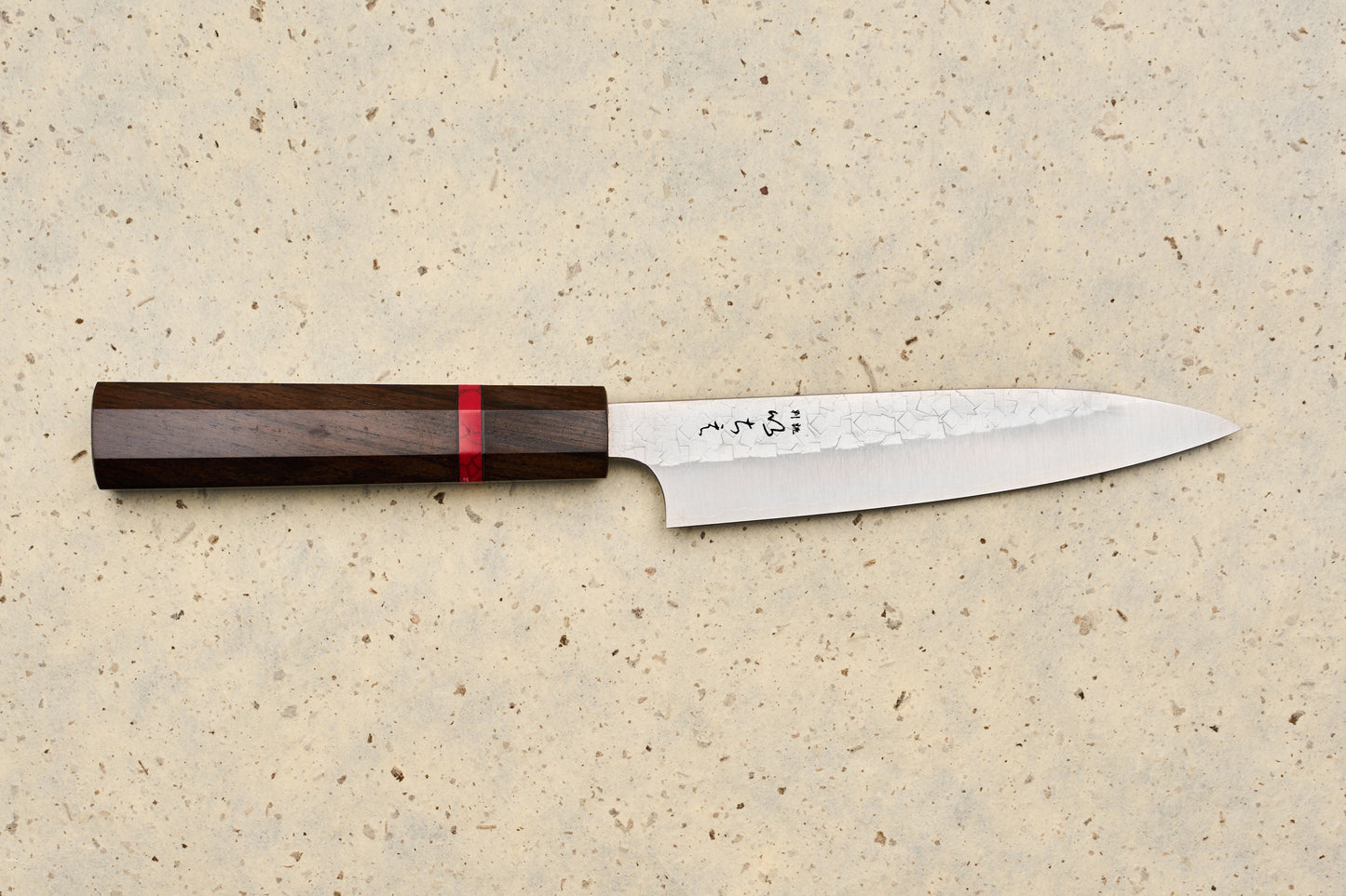 ICHIE x Shibata Knives SG2 Tsuchime Petty 135mm features a slender blade with a hammered pattern, polished edge, and dark wood handle, lying on a speckled stone surface, perfect for precise kitchen tasks.