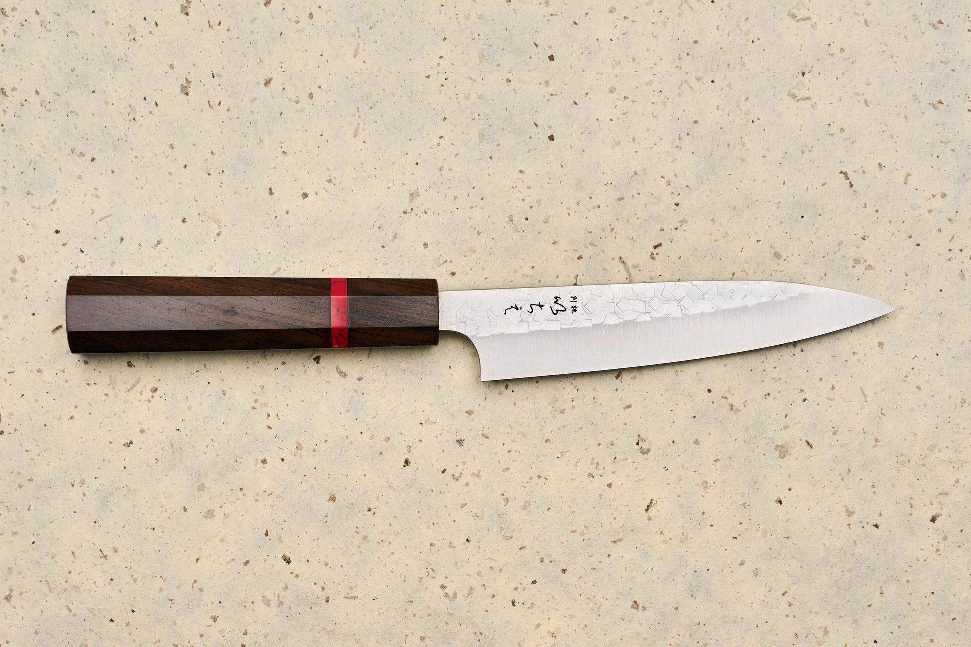 ICHIE x Shibata Knives SG2 Tsuchime Petty 135mm features a slender blade with a hammered pattern, polished edge, and dark wood handle, lying on a speckled stone surface, perfect for precise kitchen tasks.