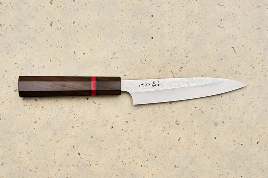 ICHIE x Shibata Knives SG2 Tsuchime Petty 135mm features a slender blade with a hammered pattern, polished edge, and dark wood handle, lying on a speckled stone surface, perfect for precise kitchen tasks.