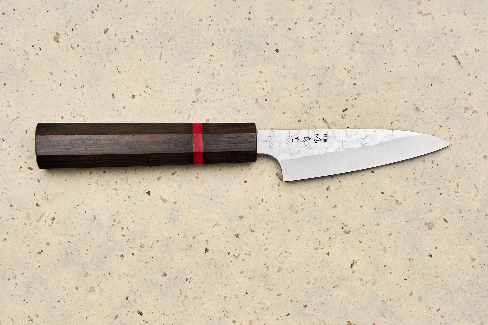 ICHIE x Shibata Knives SG2 Tsuchime Petty 90mm: A Japanese-style kitchen knife with a dark wooden octagonal handle and hammered steel blade, ideal for precision tasks on a cutting board.