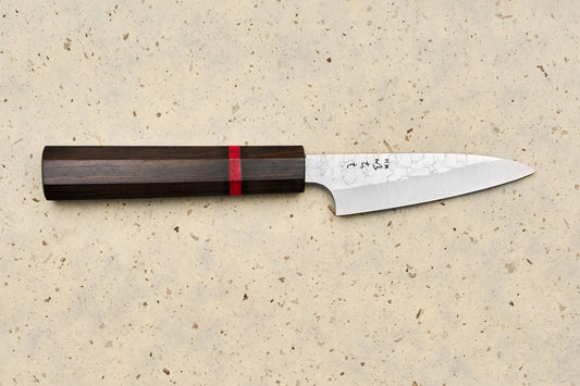 ICHIE x Shibata Knives SG2 Tsuchime Petty 90mm: A Japanese-style kitchen knife with a dark wooden octagonal handle and hammered steel blade, ideal for precision tasks on a cutting board.