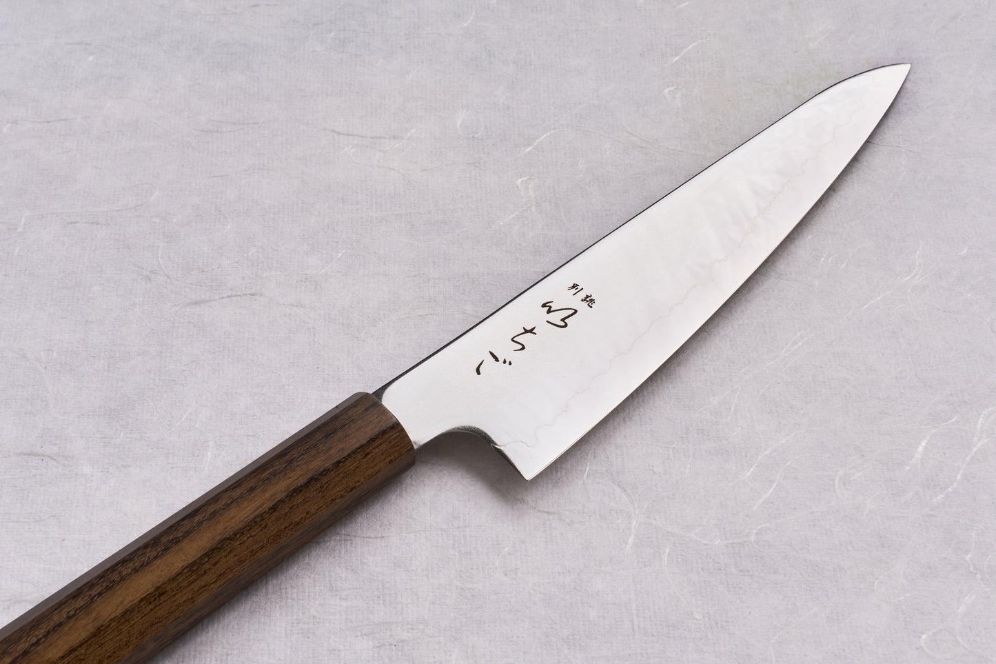 ICHIGO SLD Migaki Gyuto 150mm, a Japanese-style kitchen knife, features a polished steel blade with Japanese etchings and an octagonal wooden handle, ideal for precise cutting on a textured surface.