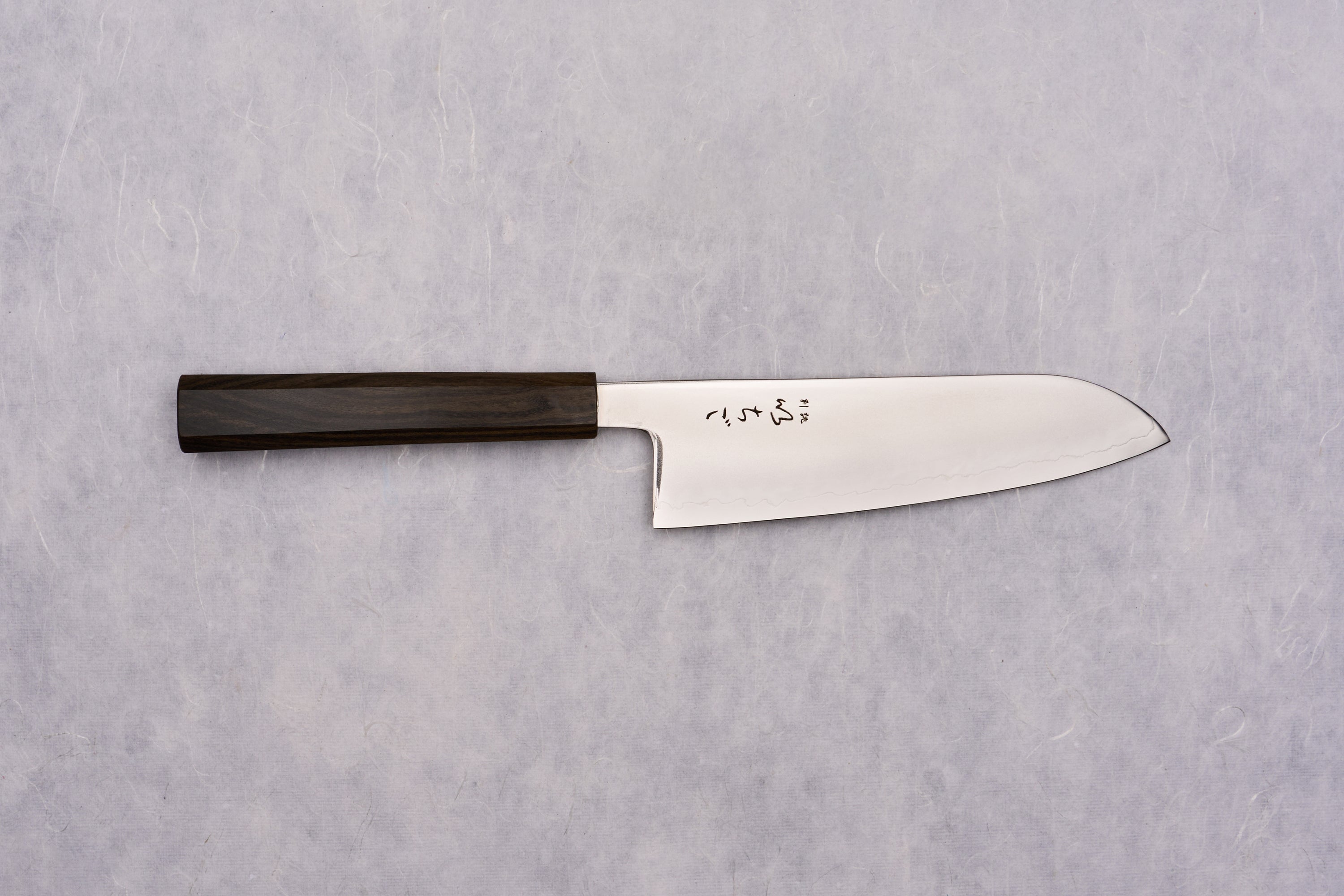 ICHIGO SLD Migaki Santoku 170mm lies flat, showcasing its polished steel blade with Japanese characters and dark wooden handle, embodying timeless craftsmanship and precision, ideal for versatile kitchen tasks.