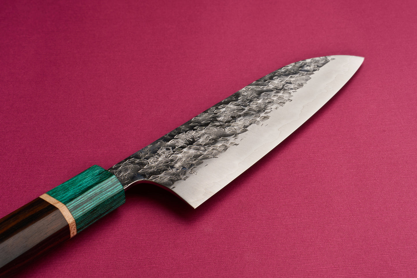 Makoto Kurosaki SPG STRIX Samidorizuki Santoku 165mm features a hammered steel blade and a layered wood handle with a green bolster, presented on reddish-pink fabric, embodying Japanese craftsmanship.