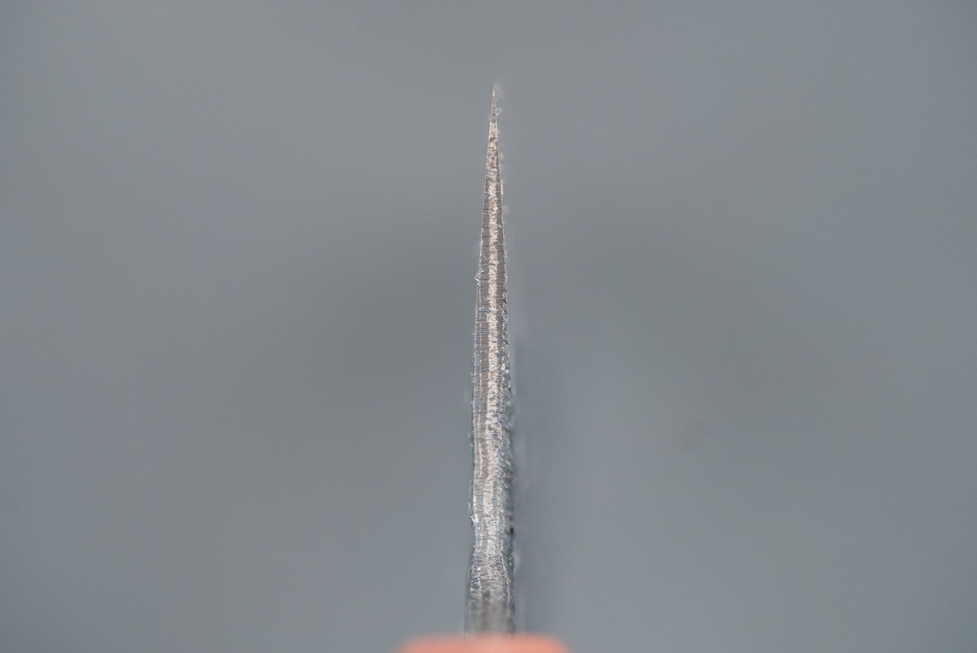Kisuke Manaka ENN Gyuto 210mm knife, showcasing its sharp, tapering Damascus-patterned blade, with a hint of an orange handle, set against a soft-focus gray background.