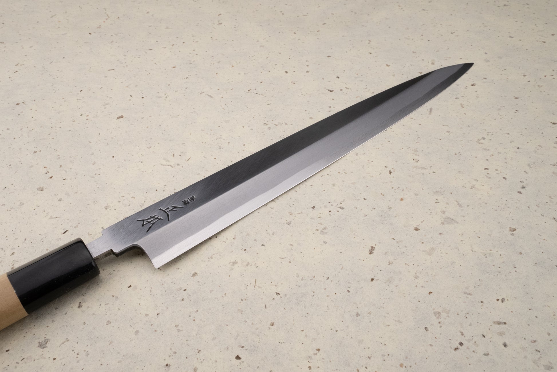 Masamoto KK Shirogami Kasumi Yanagiba 240mm knife on a speckled countertop, featuring a polished steel blade with etched Japanese characters and a traditional wooden handle with a darker bolster.