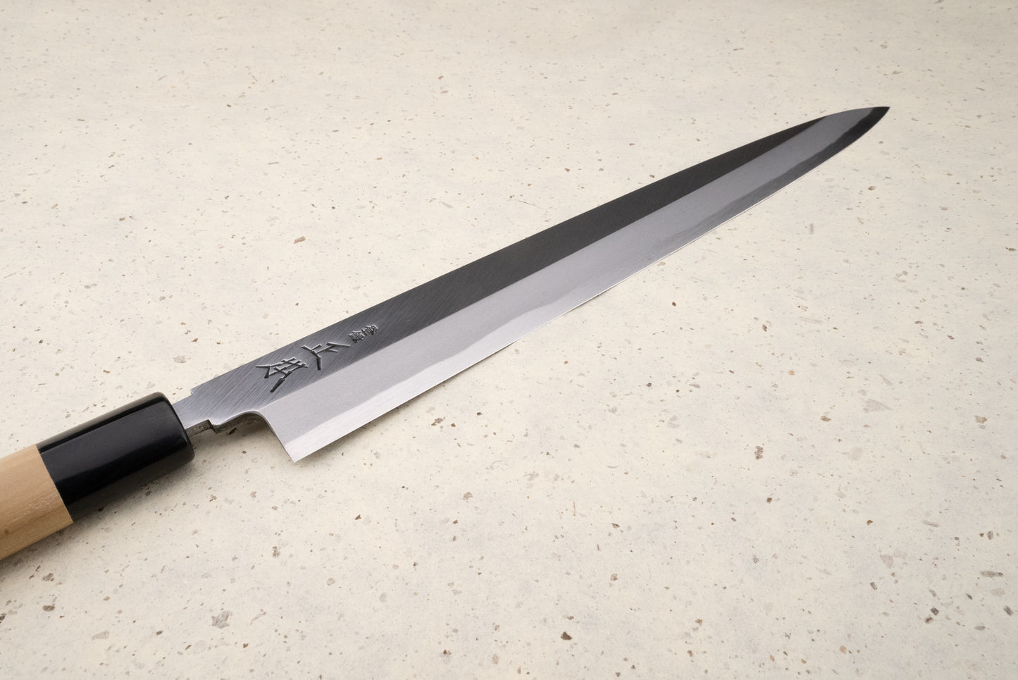 Masamoto KK Shirogami Kasumi Yanagiba 300mm, featuring a slim stainless-steel blade with Japanese engravings and a wooden handle, rests elegantly on a speckled stone surface, embodying precision and craftsmanship.
