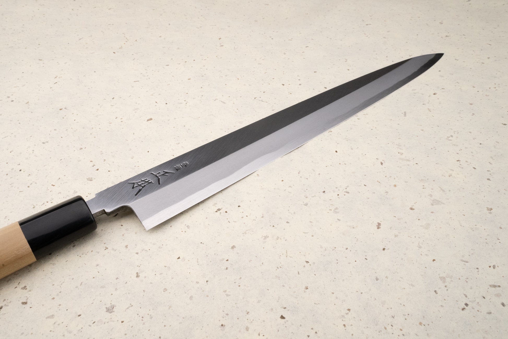 Masamoto KK Shirogami Kasumi Yanagiba 300mm, featuring a slim stainless-steel blade with Japanese engravings and a wooden handle, rests elegantly on a speckled stone surface, embodying precision and craftsmanship.