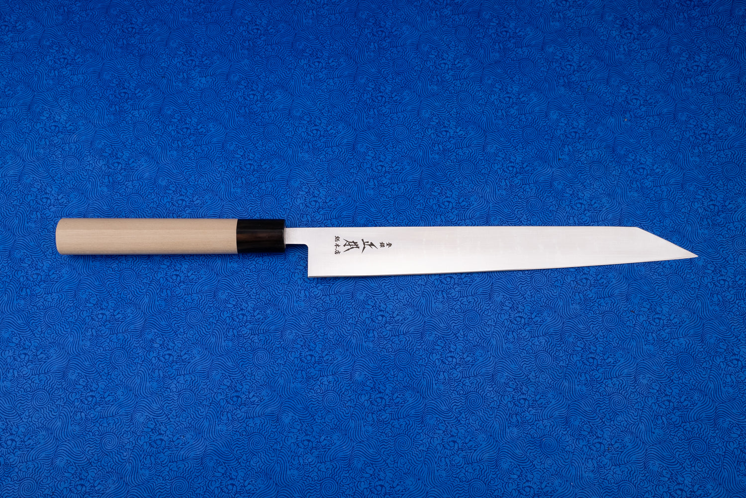 Masamoto MV Kensaki Sujihiki 255mm knife displayed on a blue patterned cloth, showcasing its sharp, narrow steel blade with Japanese etchings, black ferrule, and light wood handle, ideal for precise slicing.