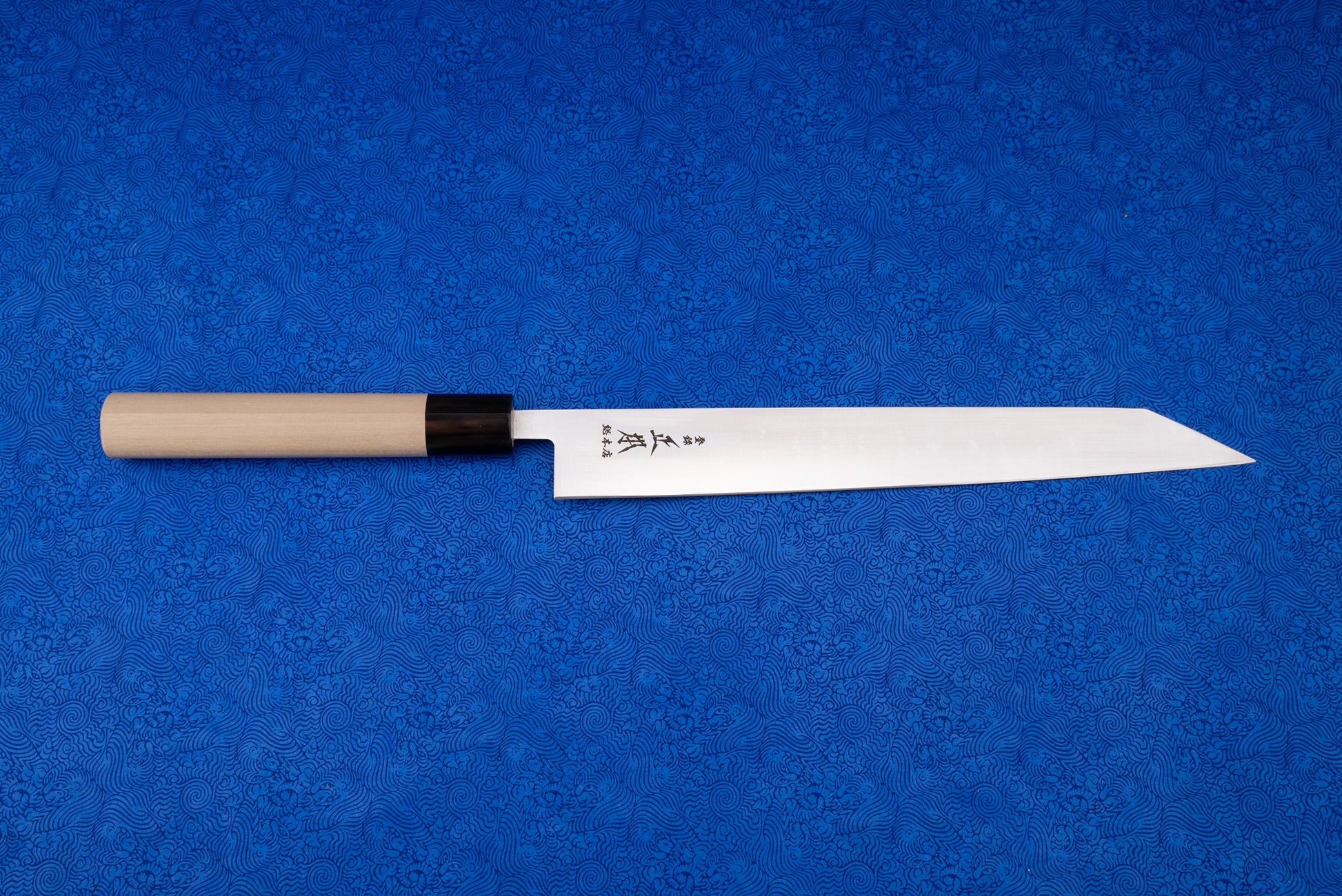Masamoto MV Kensaki Sujihiki 255mm knife displayed on a blue patterned cloth, showcasing its sharp, narrow steel blade with Japanese etchings, black ferrule, and light wood handle, ideal for precise slicing.