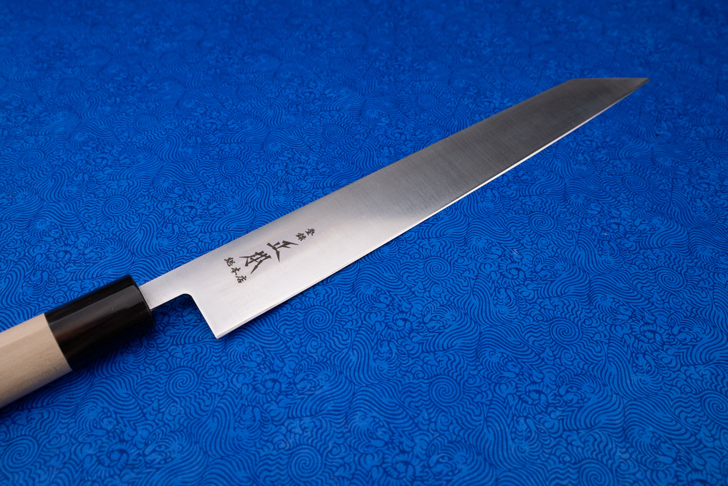 Masamoto MV Kensaki Sujihiki 255mm, a sleek Japanese kitchen knife with a wooden handle and engraved stainless blade, displayed on a blue cloth, exemplifies precision and craftsmanship for slicing meats and fish.