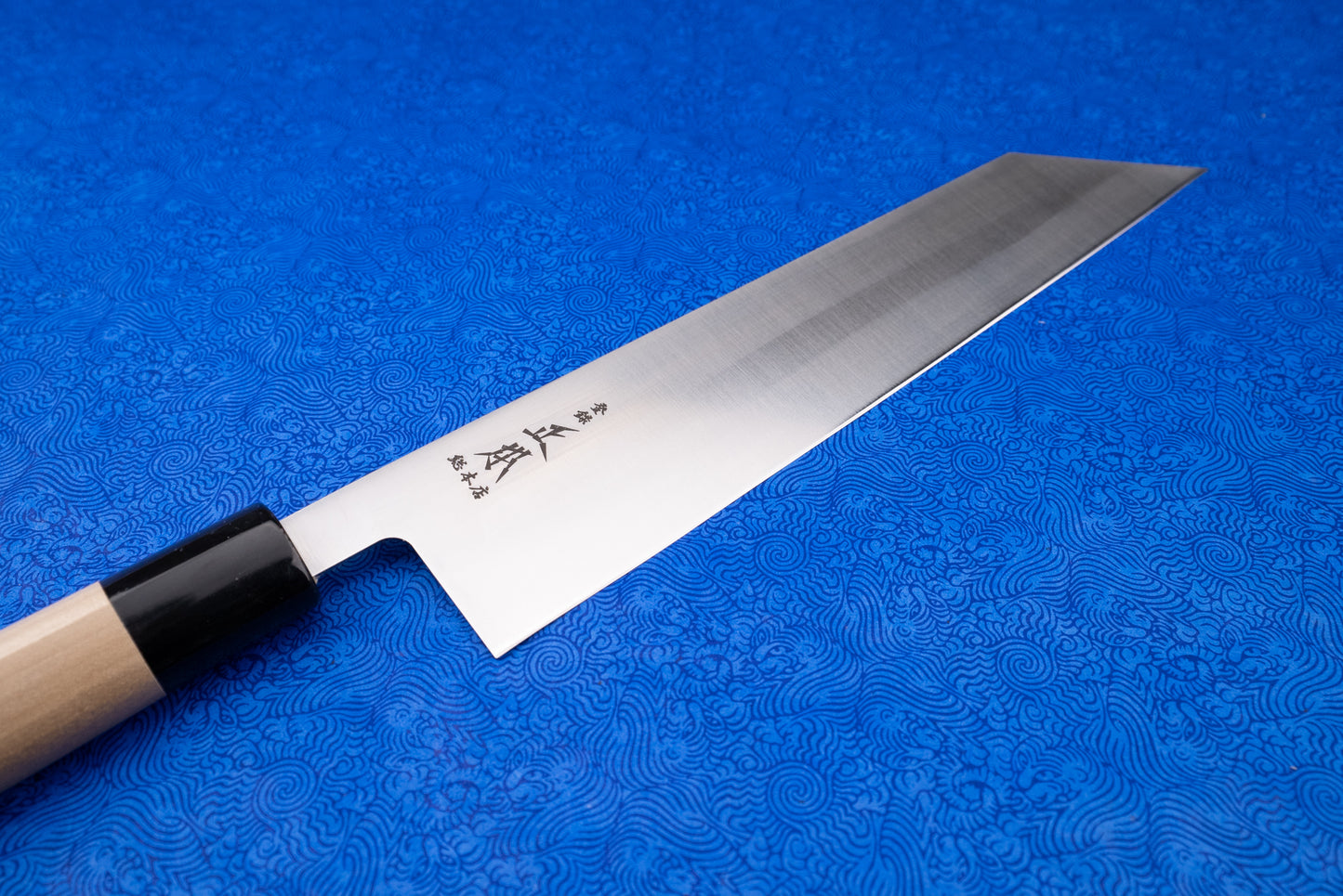 Masamoto MV Kiritsuke Gyuto 240mm knife with a long, thin single-bevel blade featuring Japanese characters, a black ferrule, and a simple wooden handle displayed on a blue, patterned cloth.