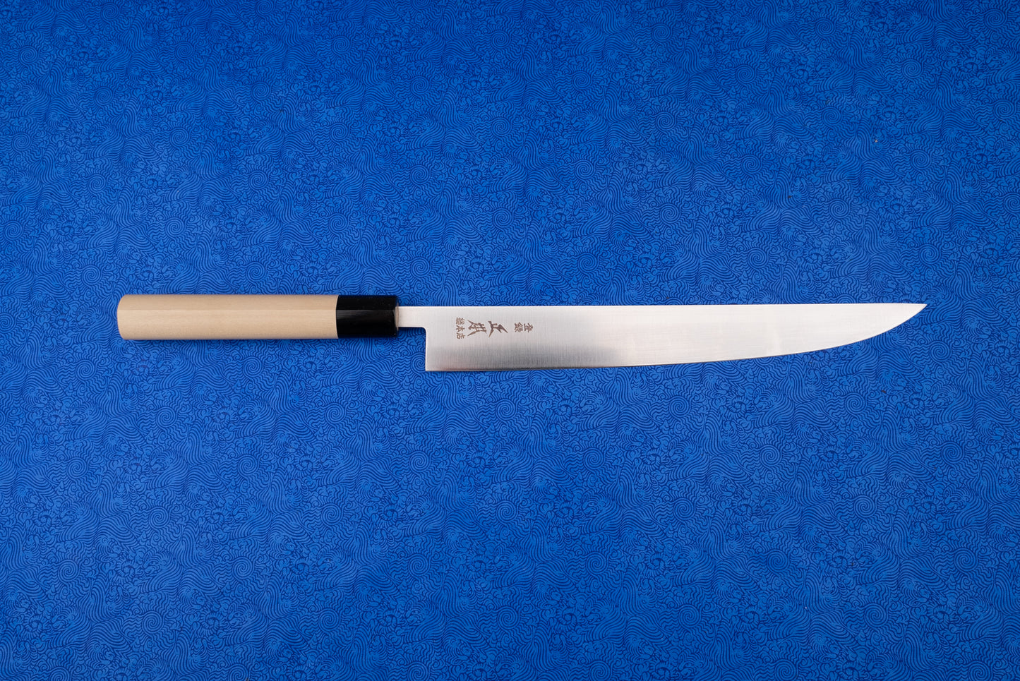 Masamoto MV Tachigata Sujihiki 255mm, a Japanese-style slicing knife, features a single-bevel stainless steel blade with kanji and a natural wood handle, displayed on a vibrant blue patterned cloth.