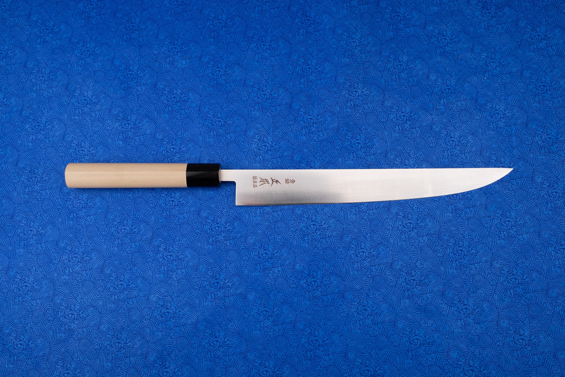 Masamoto MV Tachigata Sujihiki 255mm, a Japanese-style slicing knife, features a single-bevel stainless steel blade with kanji and a natural wood handle, displayed on a vibrant blue patterned cloth.