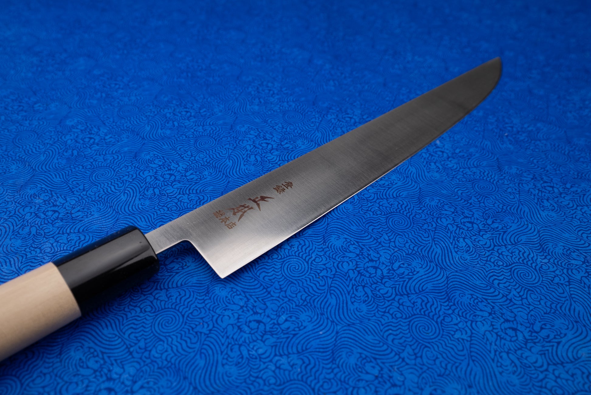Masamoto MV Tachigata Sujihiki 255mm, a single-bevel Japanese slicing knife, features a slender steel blade with kanji and an octagonal wooden handle, resting on a blue wave-patterned cloth.