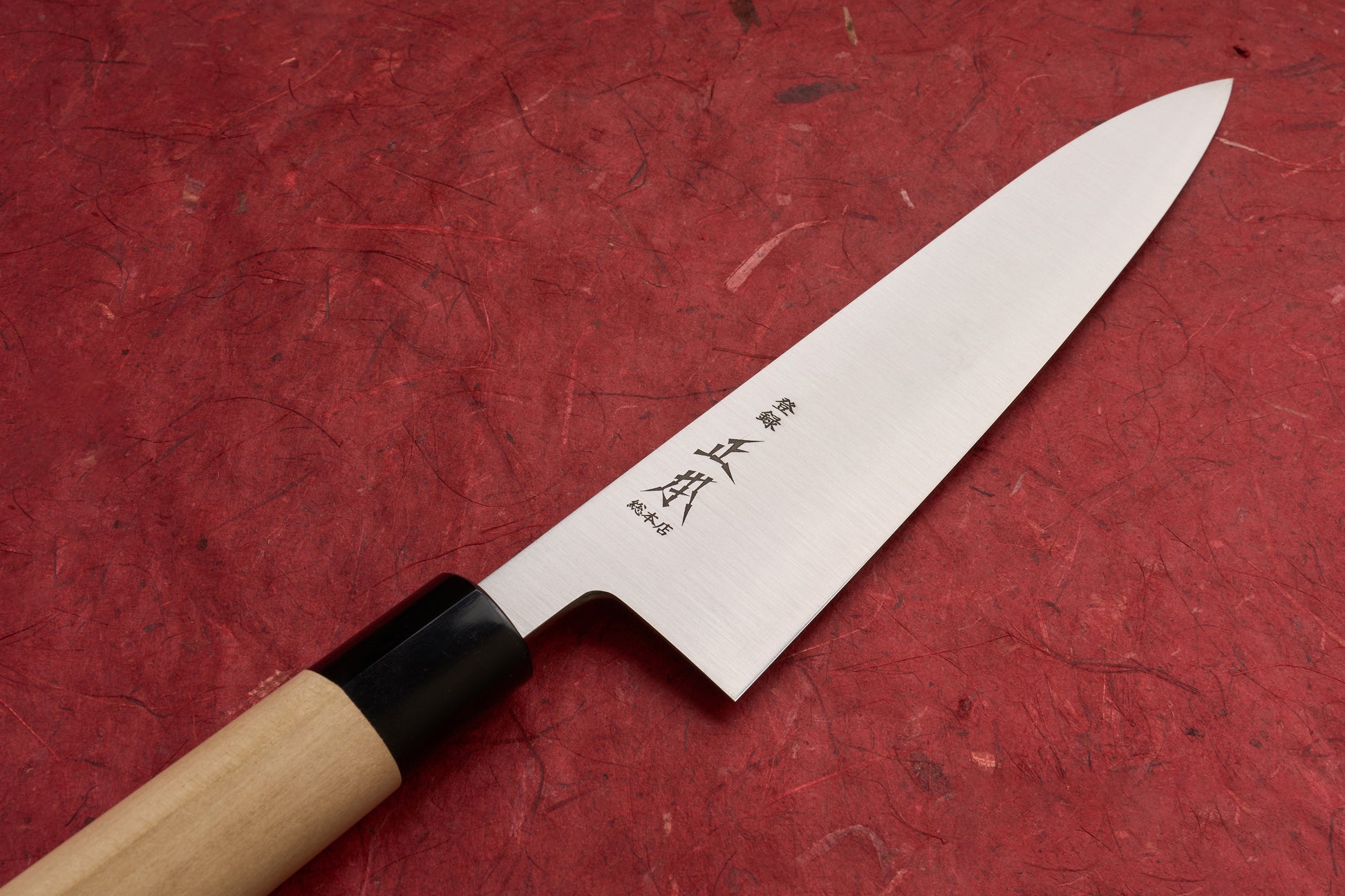 Masamoto KS Hongasumi Gyuto 210mm knife with a stainless-steel blade engraved with Japanese characters, resting on a deep red textured background, showcasing its sleek design and wooden handle with black bolster.