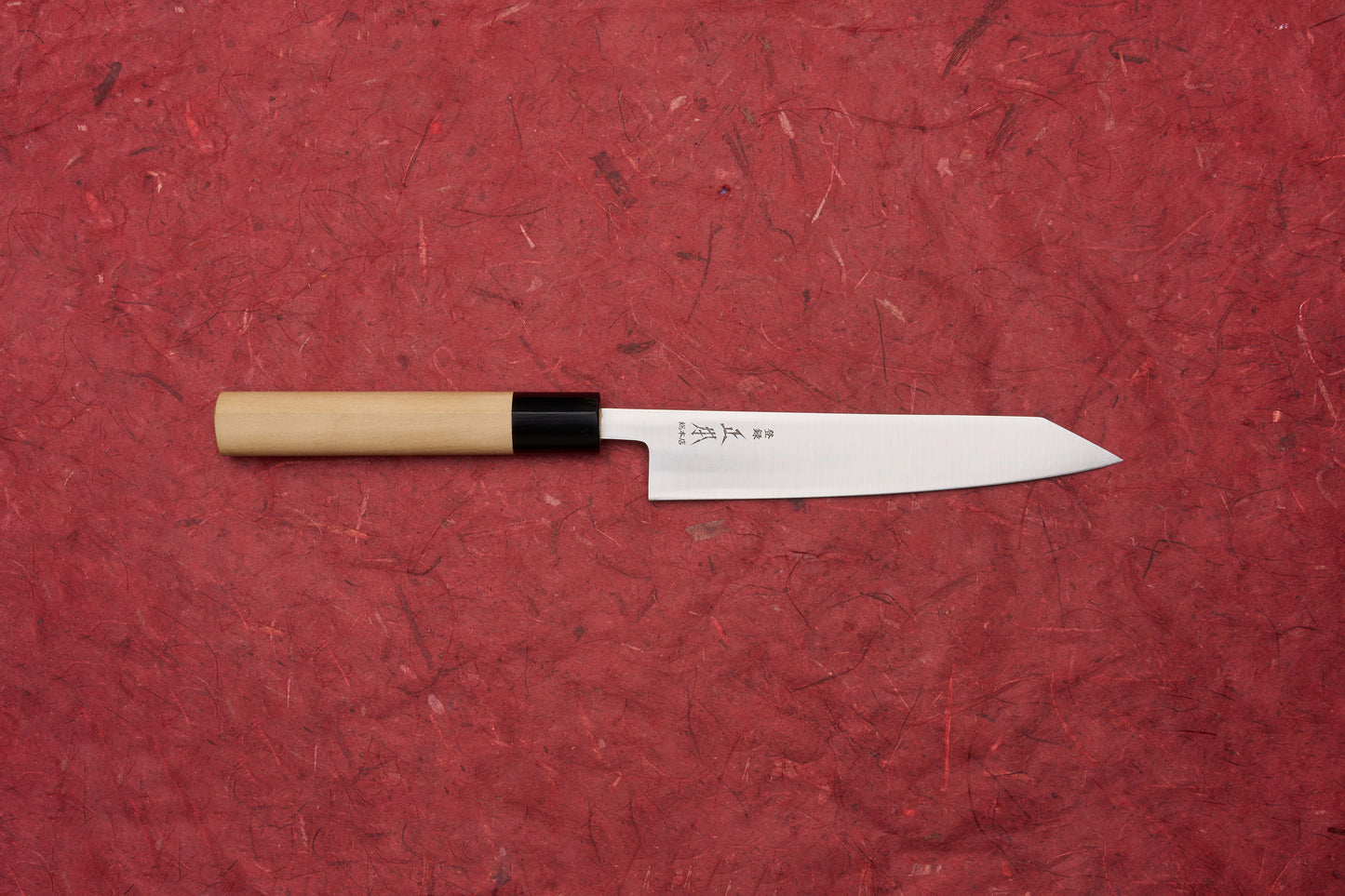 Masamoto KS Hongasumi Kiritsuke Petty 165mm knife with a polished steel blade and Japanese characters, featuring a light wood handle, showcased on a red textured surface.