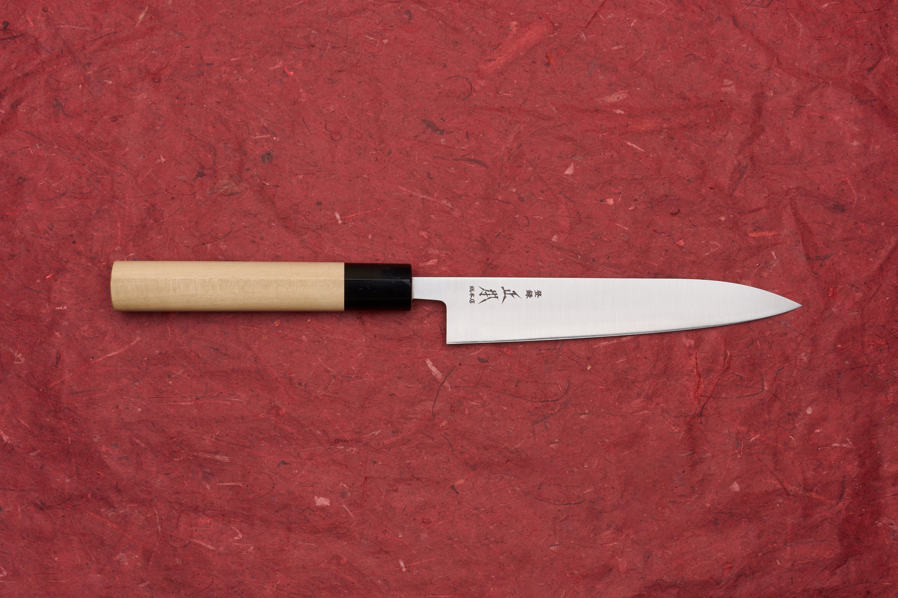 Masamoto KS Hongasumi Petty 165mm, a meticulously crafted Japanese kitchen knife with a polished steel blade and light wooden handle, displayed on a red surface, exemplifying Masamoto's renowned precision and thinness.
