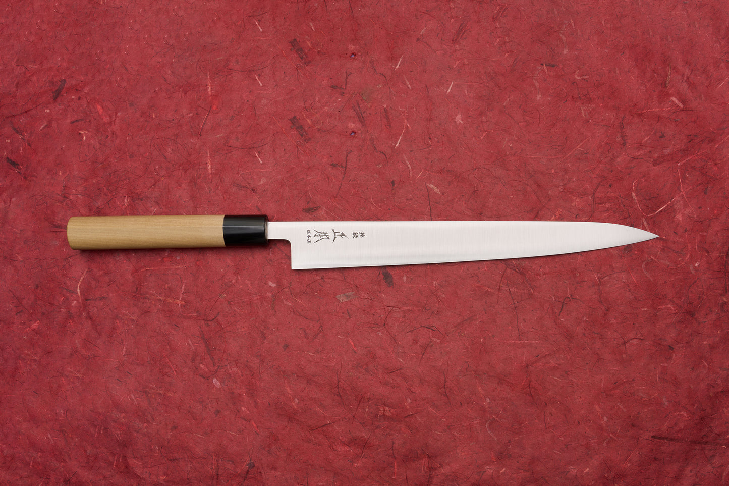 Masamoto KS Hongasumi Sujihiki 270mm laid horizontally on a red surface, showcasing its slender, sharp blade with kanji markings and a traditional wooden handle, epitomizing Japanese craftsmanship.