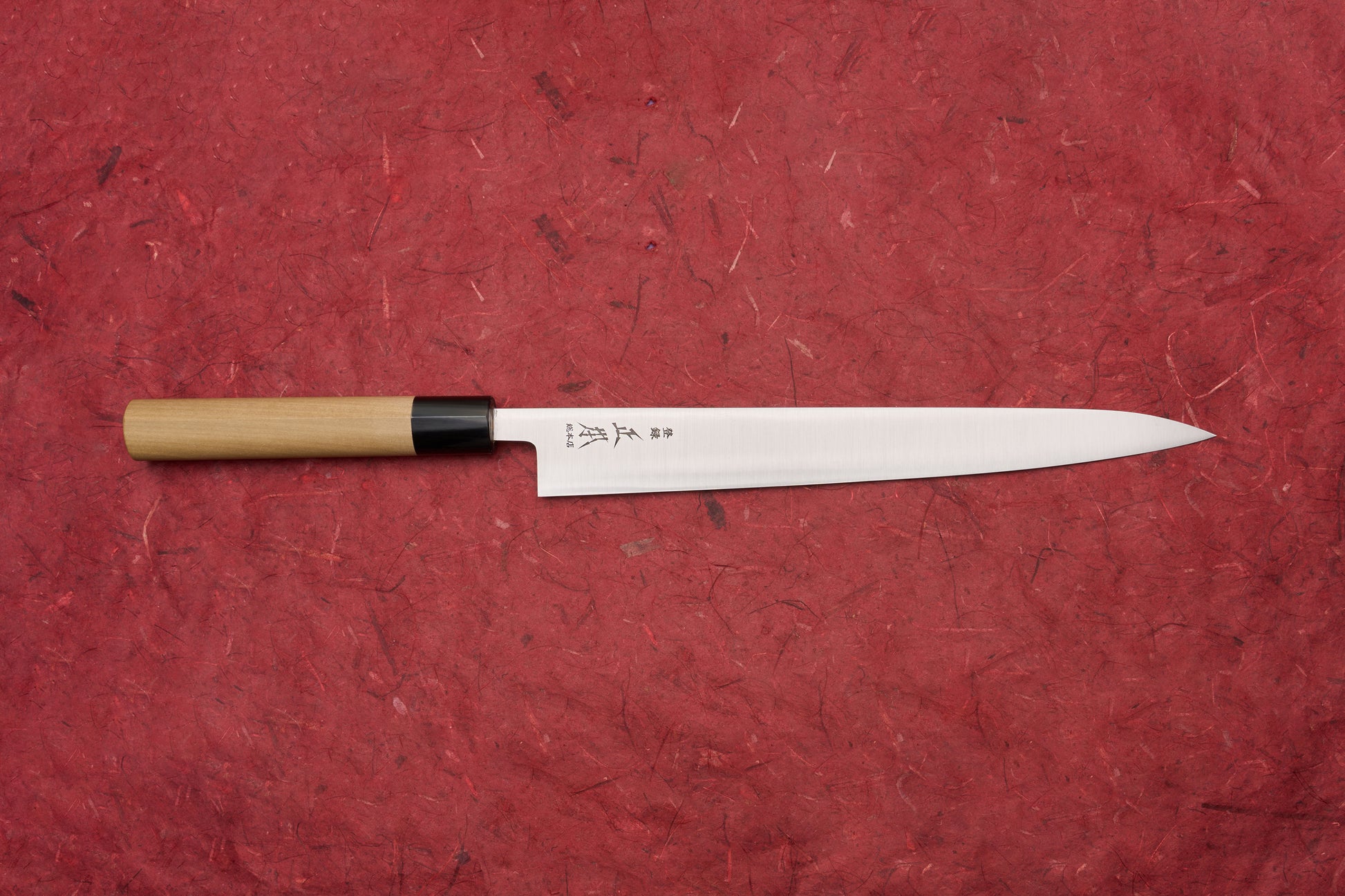 Masamoto KS Hongasumi Sujihiki 270mm laid horizontally on a red surface, showcasing its slender, sharp blade with kanji markings and a traditional wooden handle, epitomizing Japanese craftsmanship.