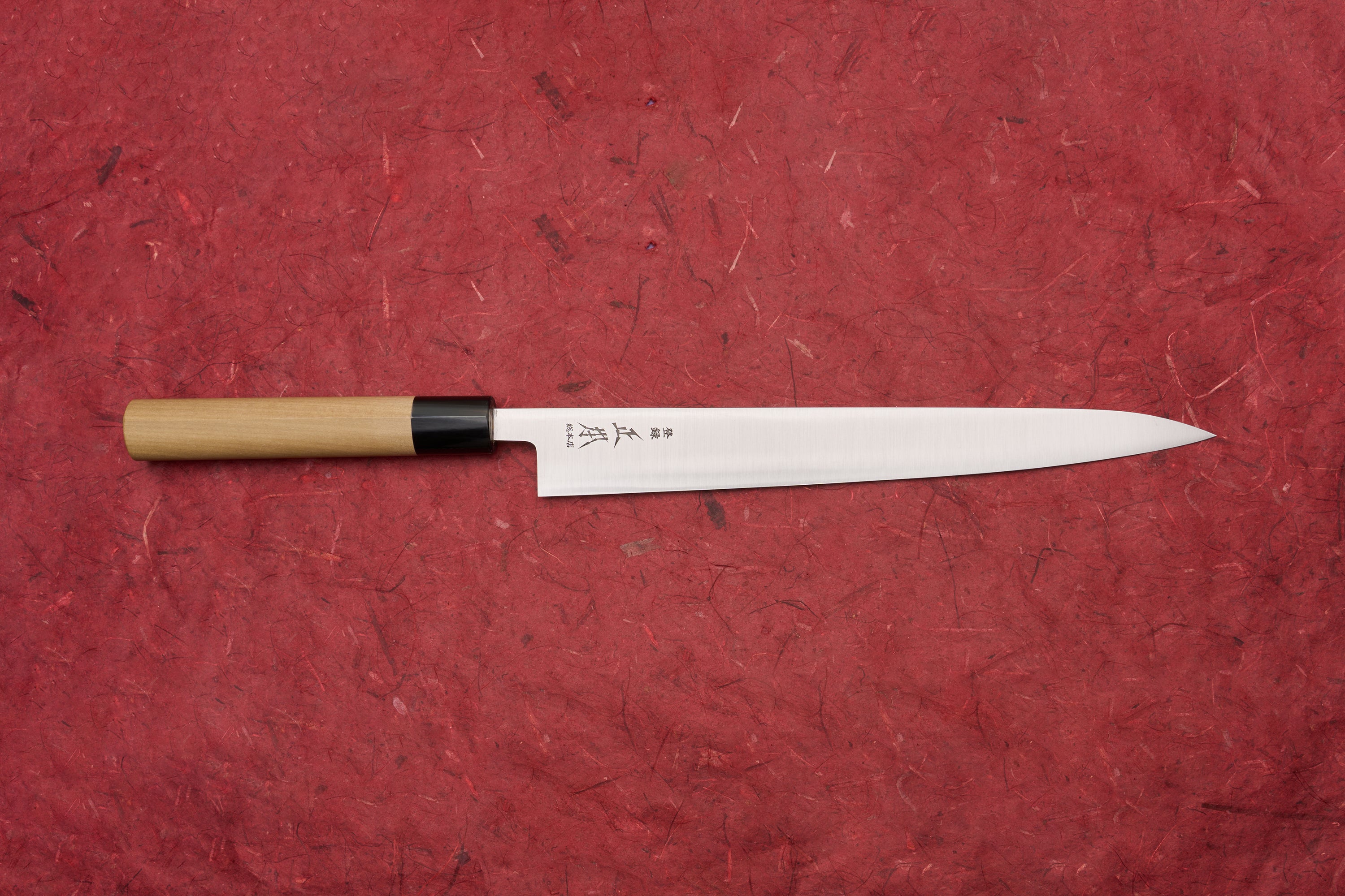 Masamoto KS Hongasumi Sujihiki 270mm laid horizontally on a red surface, showcasing its slender, sharp blade with kanji markings and a traditional wooden handle, epitomizing Japanese craftsmanship.